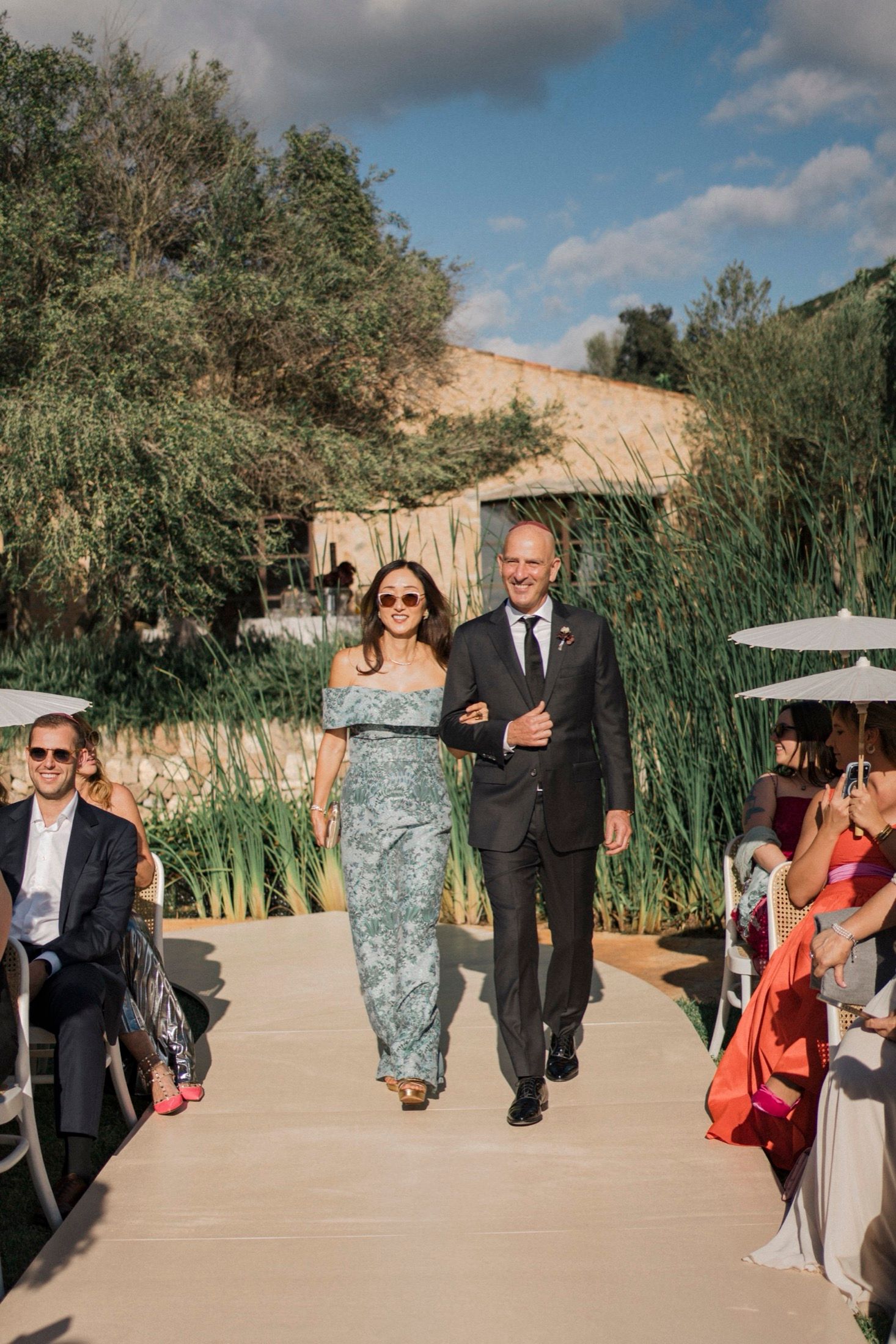 Wedding party members walk down an outdoor pathway lined with tall reeds and desert landscaping under blue skies.