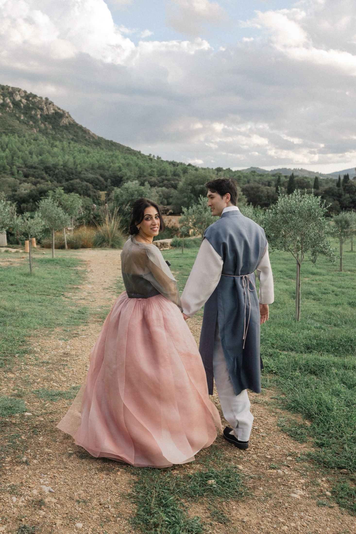 A couple walks together in a scenic mountain landscape with one wearing a flowing pink dress and the other in blue attire.