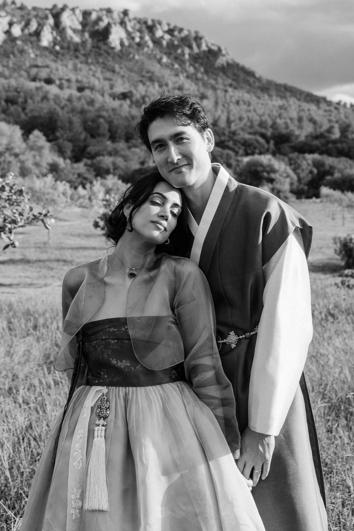 A couple in traditional Korean hanbok attire pose together in a scenic outdoor mountain setting.