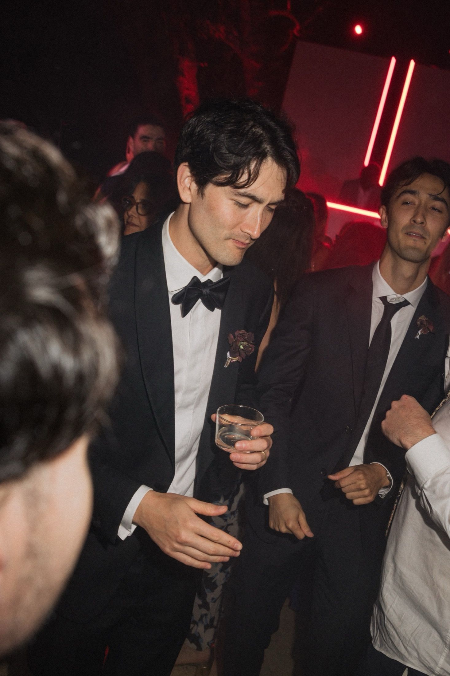 A group of people in formal attire socializing at a dimly lit party with red lighting and dark ambiance.