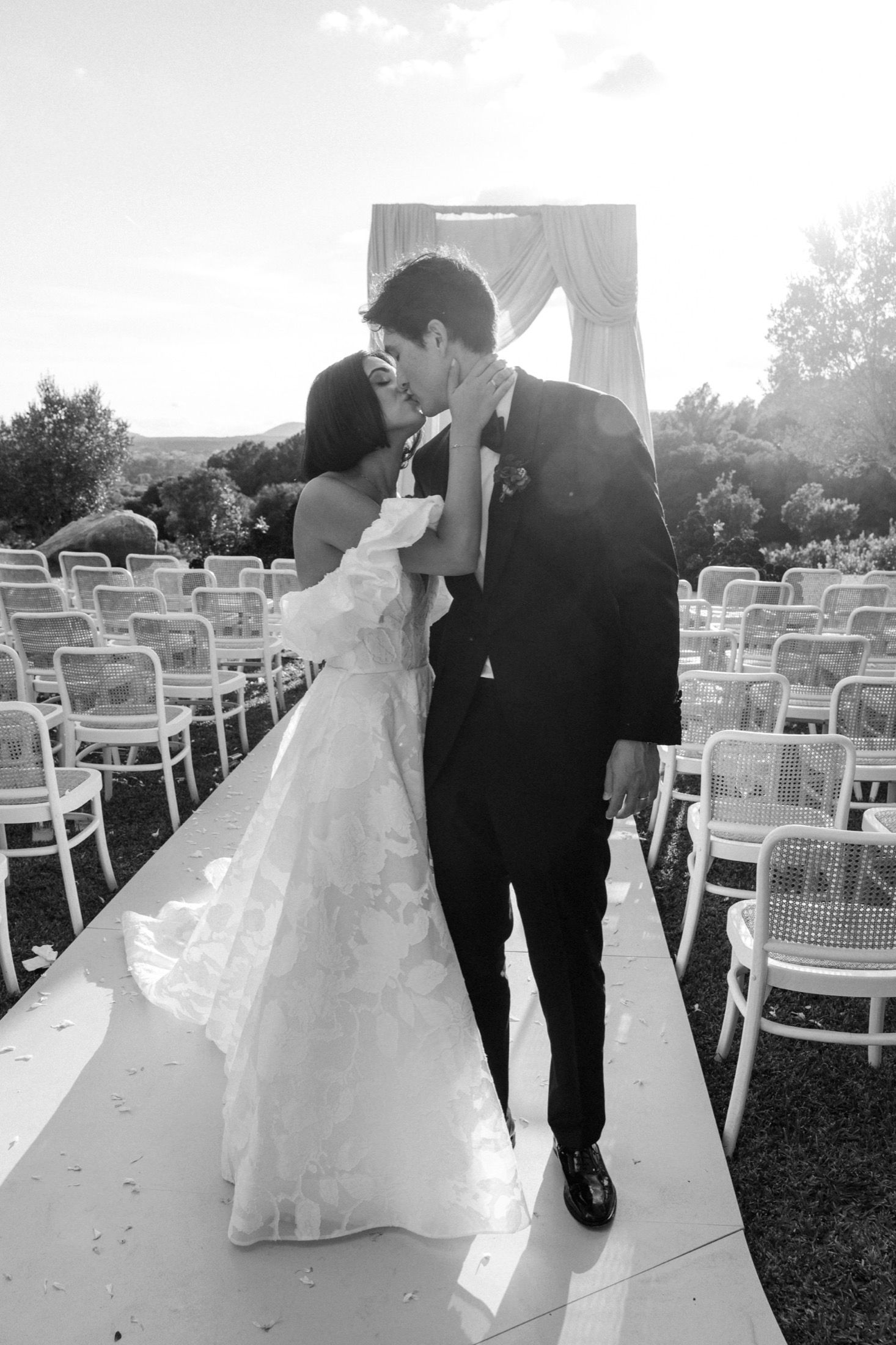 A bride and groom walk away together through rows of white garden chairs at an outdoor wedding ceremony.