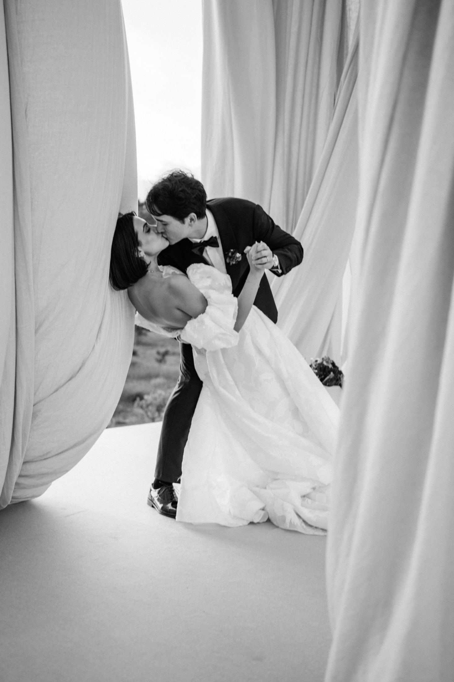 A black and white wedding photo of a romantic dip between flowing white curtains outdoors.