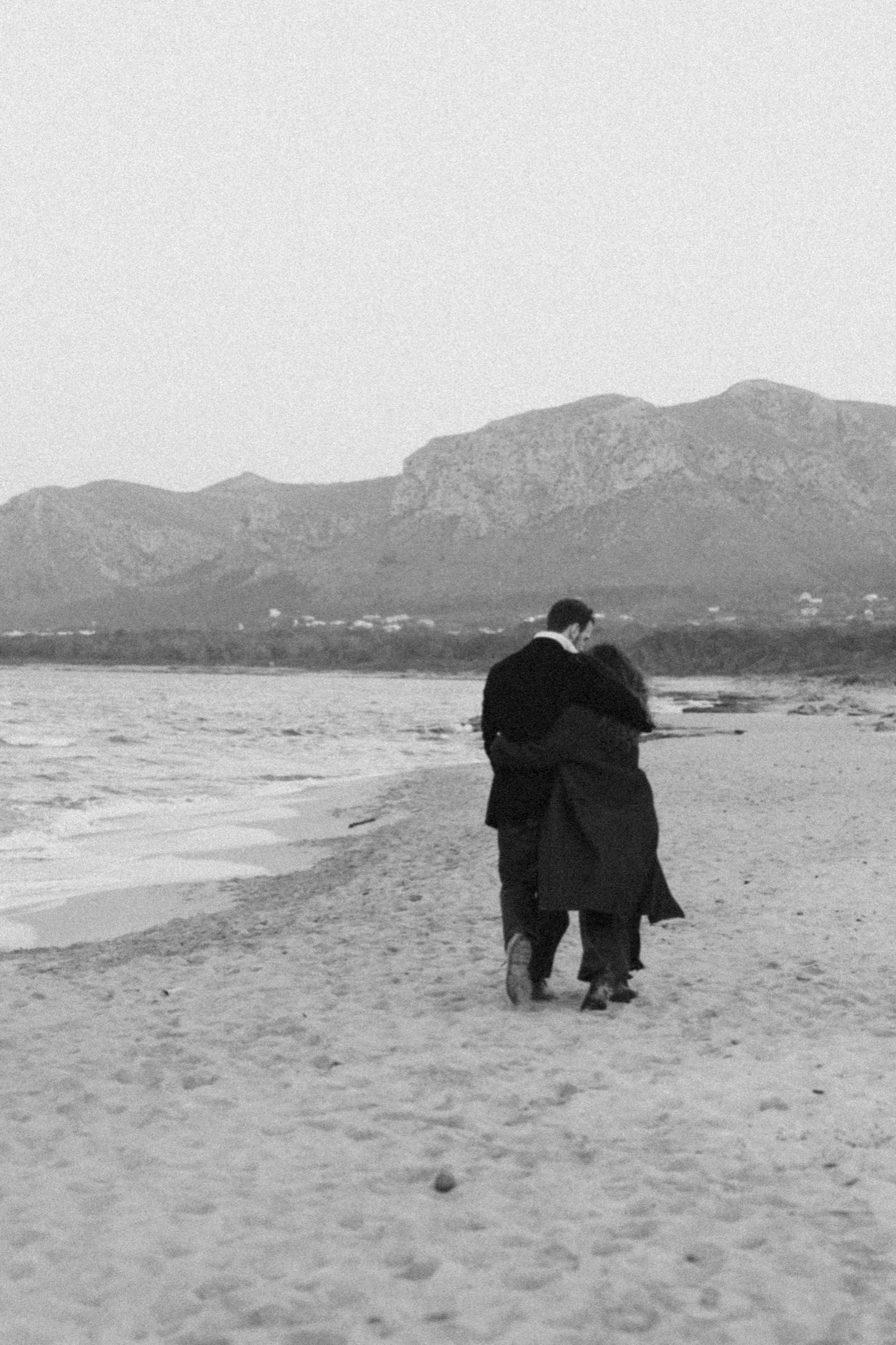Two figures in dark clothing walk along a serene beach with mountains in the distance, captured in black and white.