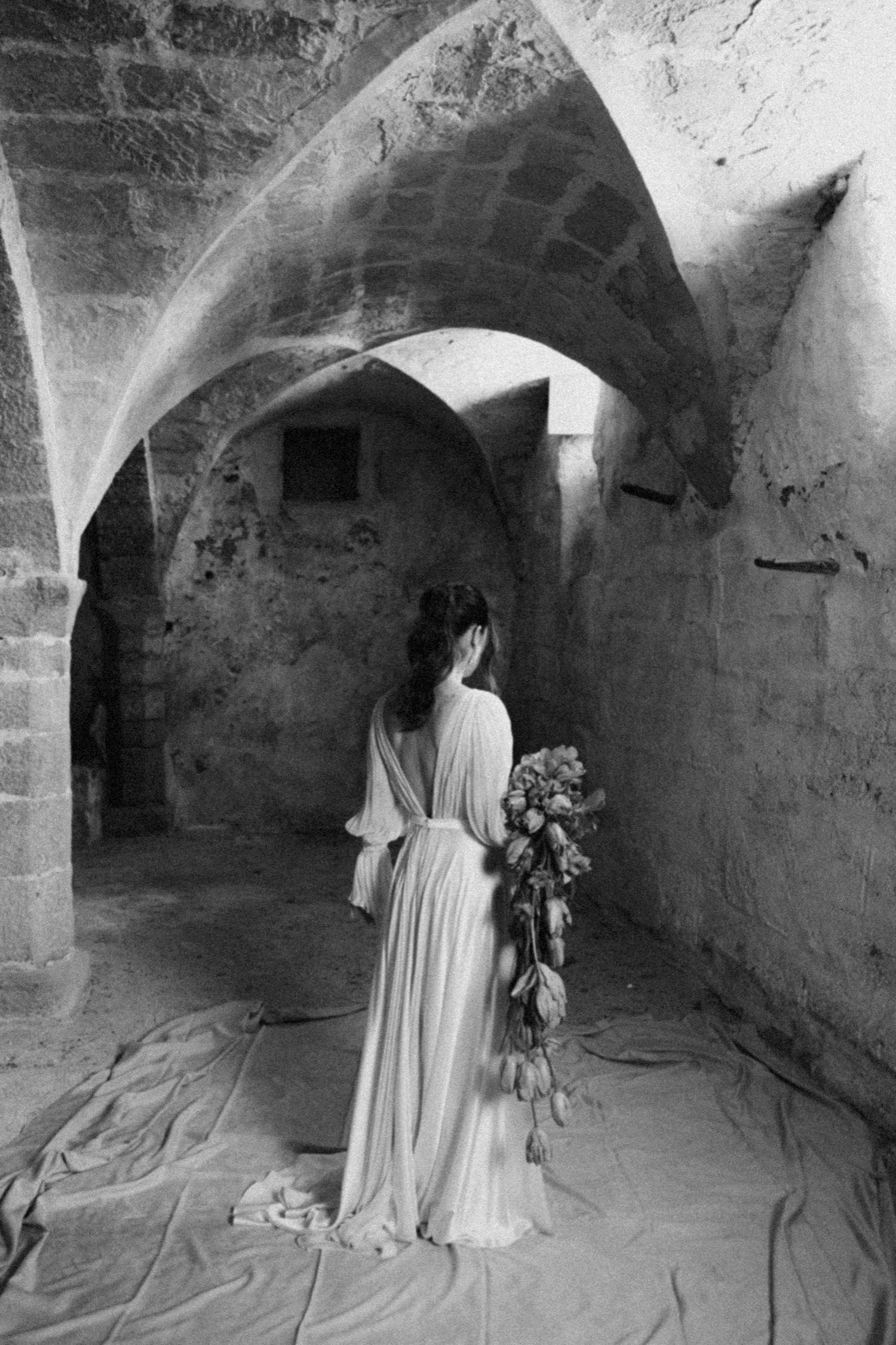 A ghostly figure in white stands in a dark stone tunnel holding flowers in this haunting black and white photograph series.
