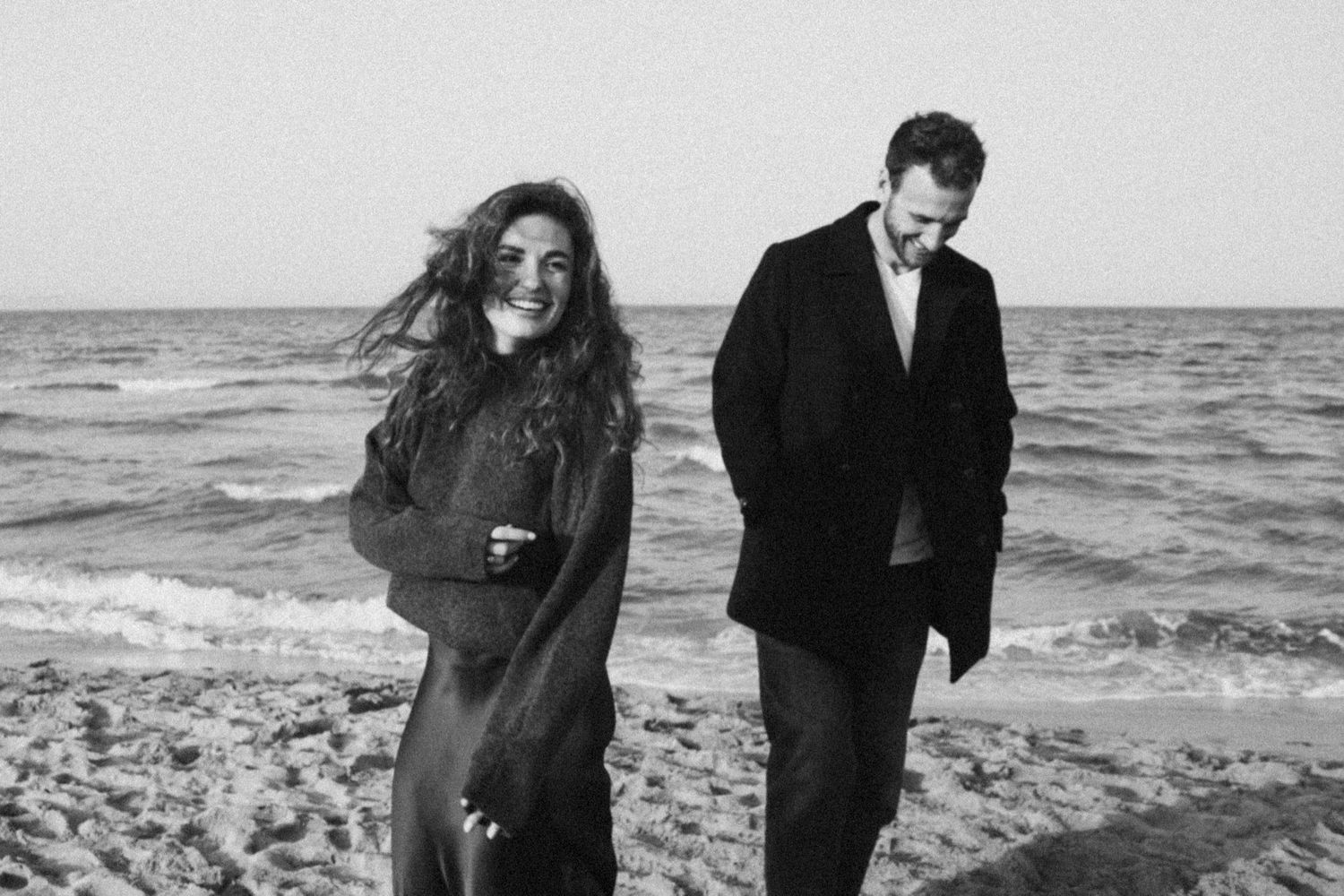 A couple shares a romantic moment walking along a windy beach during a black and white photoshoot.