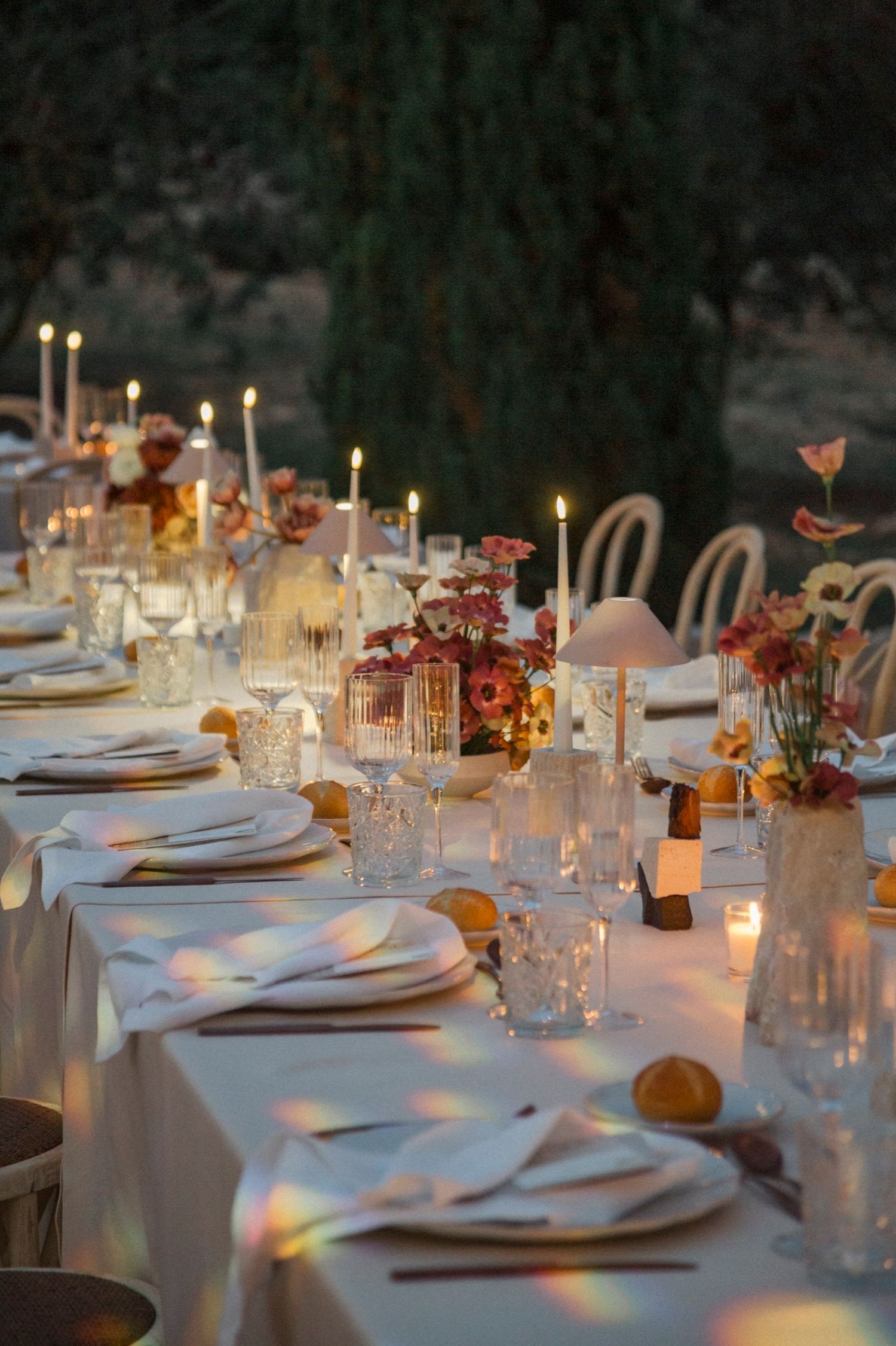 Elegant dinner table setting with lit candles, white linens, and rainbow light reflections creating a romantic ambiance.