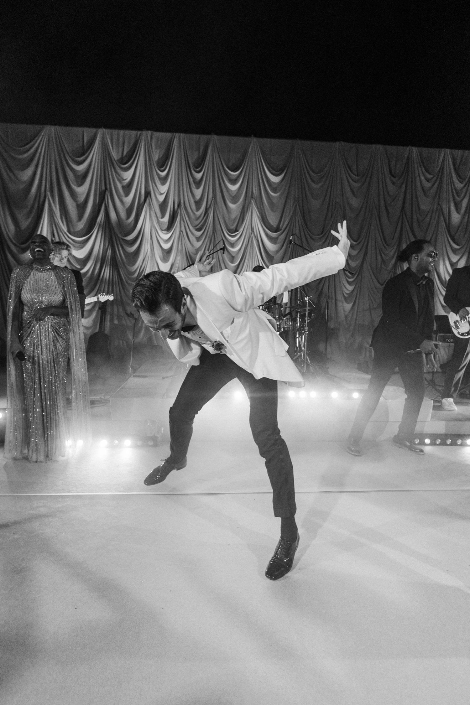 A performer in a white jacket dances energetically on a stage with dramatic curtains and fog effects in black and white.