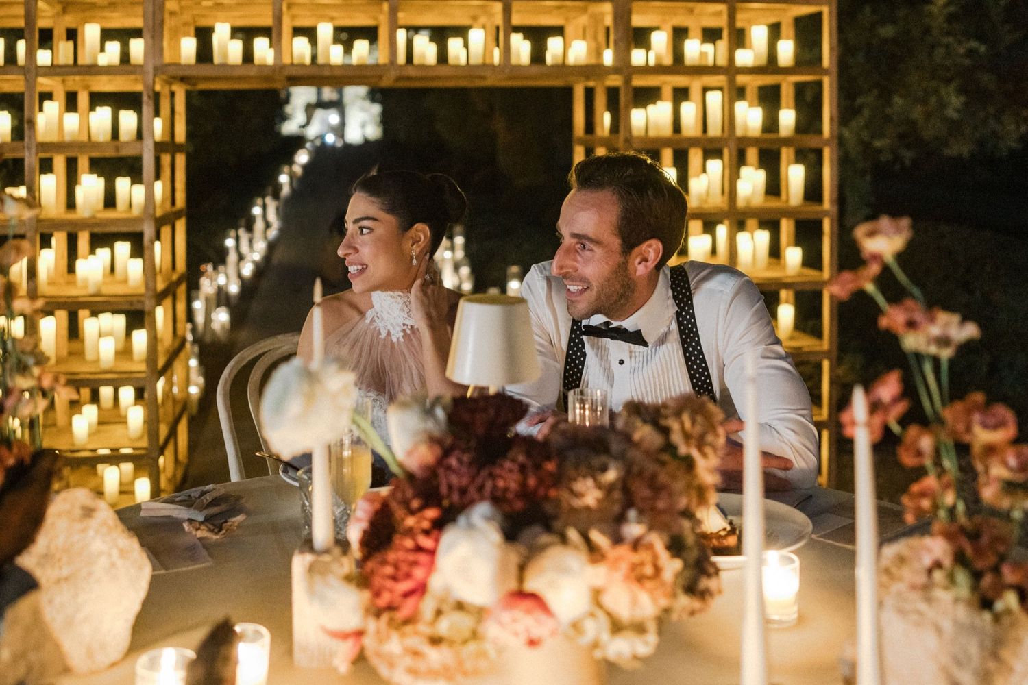 Wedding reception guests seated at long tables decorated with candles and floral arrangements in a rustic venue.