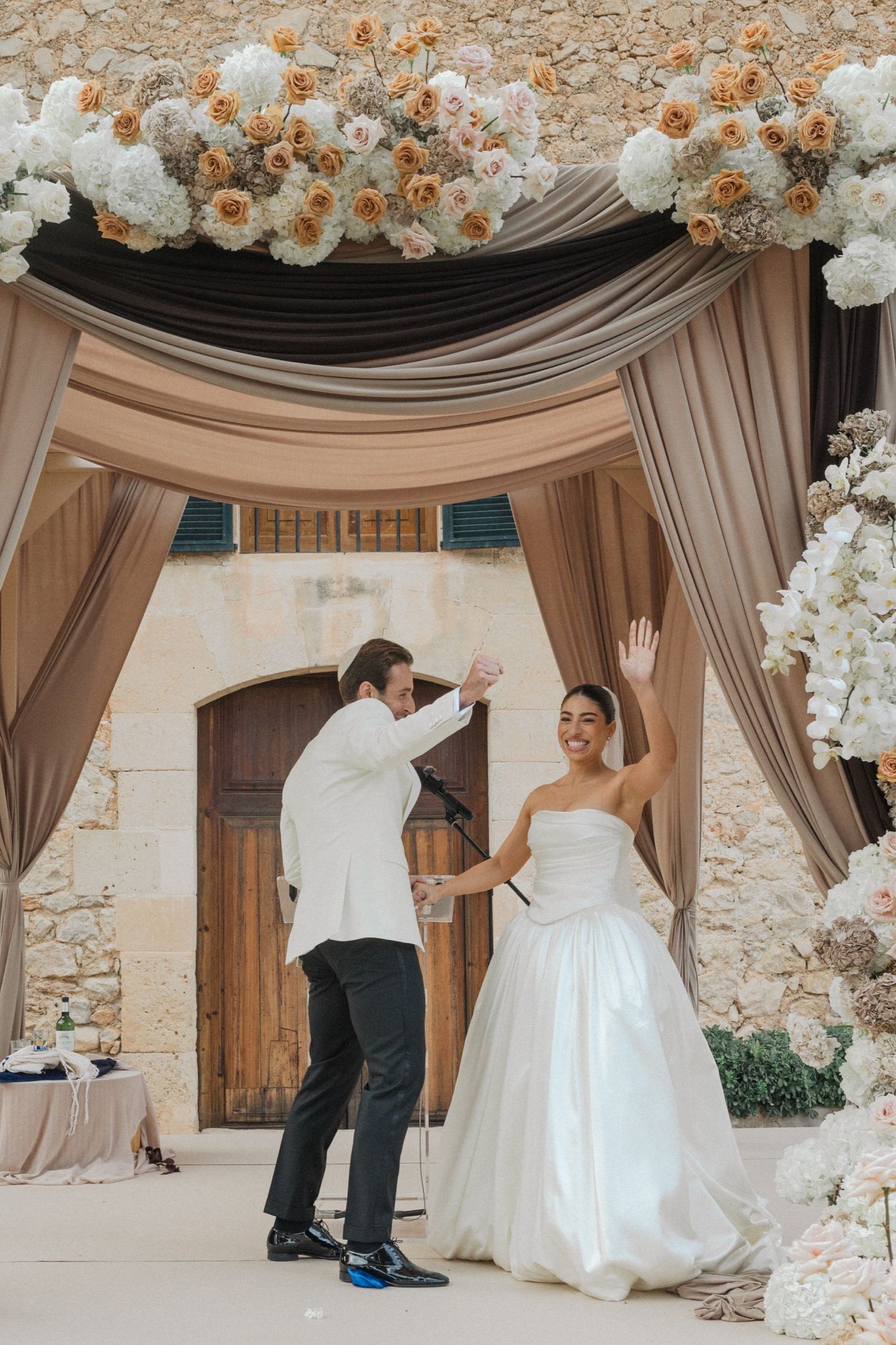 A joyful wedding celebration under an elegant draped arch decorated with white and blush floral arrangements.