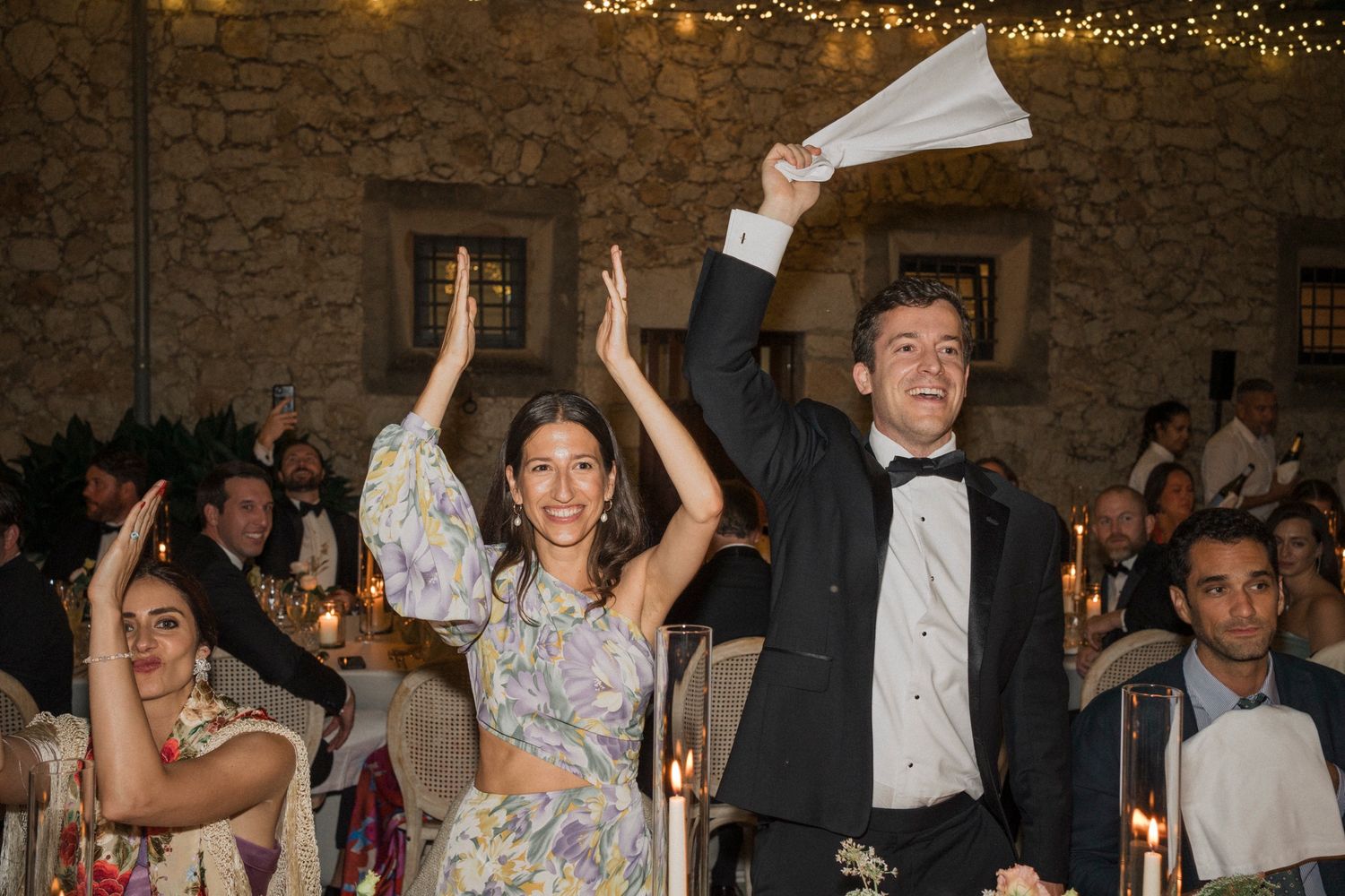 Wedding guests celebrate joyfully with raised arms during a reception in a rustic stone venue with twinkling lights.