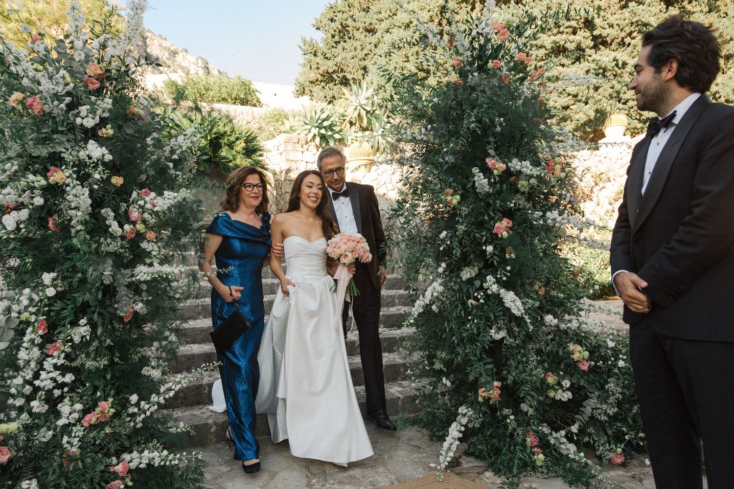 A romantic garden wedding ceremony takes place under a lush floral arch with climbing roses and greenery.