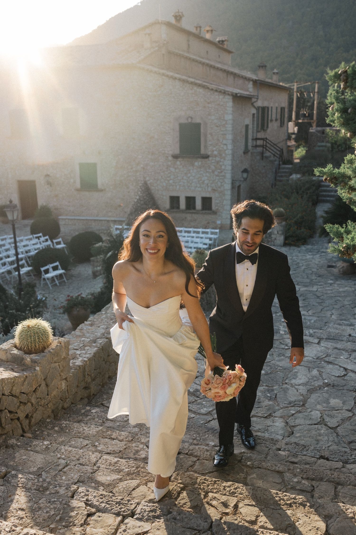 A couple in formal wedding attire walks along a rustic stone path on a sunny afternoon.