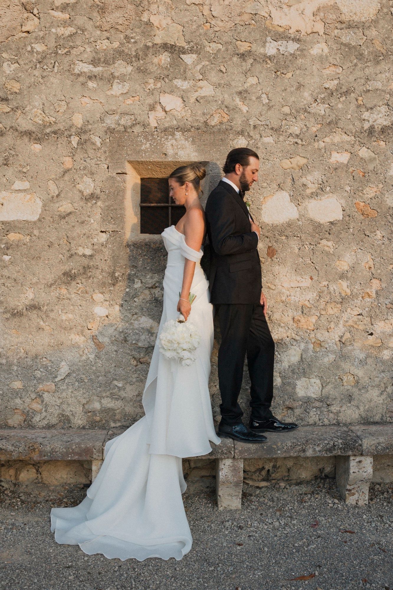Newlyweds pose for creative wedding portraits along a historic stone wall with gravel ground.