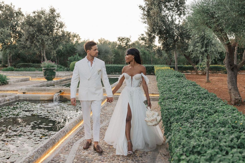 Wedding couple walking through a lush garden path in white attire.