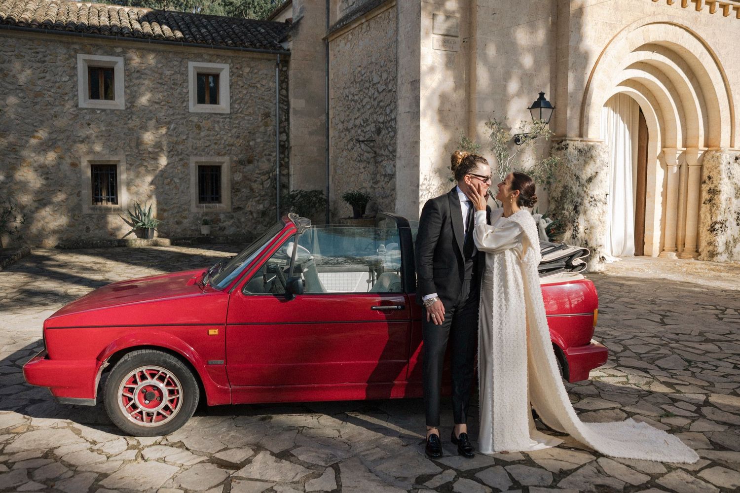 Wedding photos with red vintage car and elegant white dress against historic stone architecture.