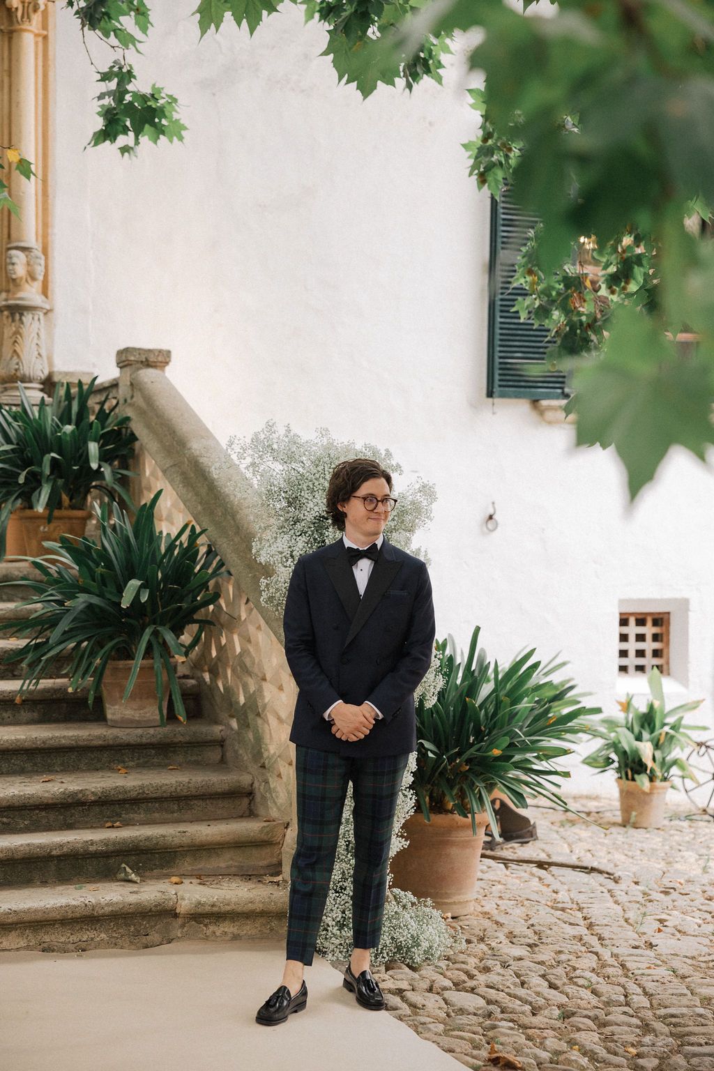 A groom in a black tuxedo and bow tie stands on stone steps against a white wall with plants.