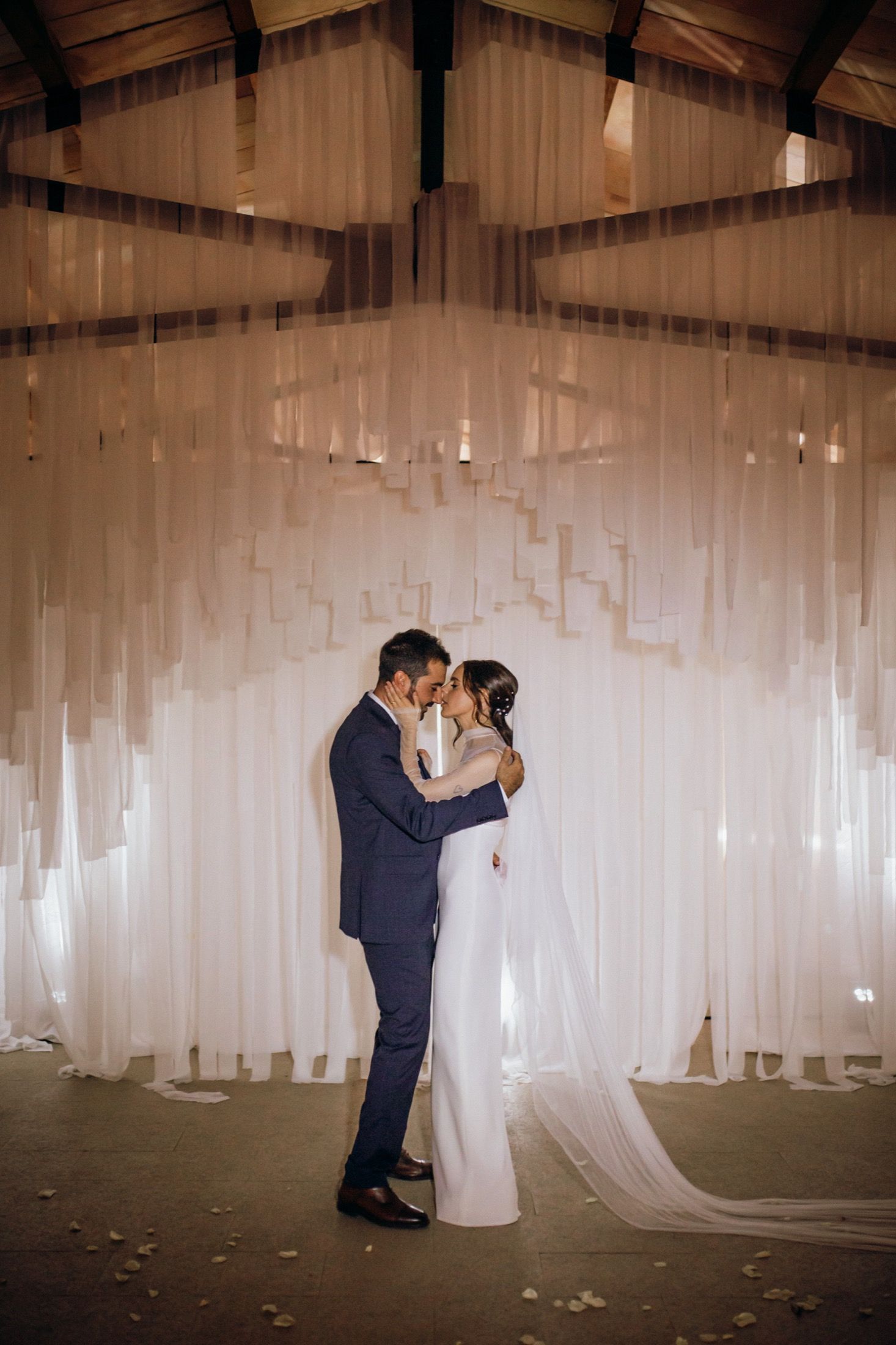 A couple shares a romantic dance moment surrounded by flowing white fabric panels in a dimly lit reception venue.