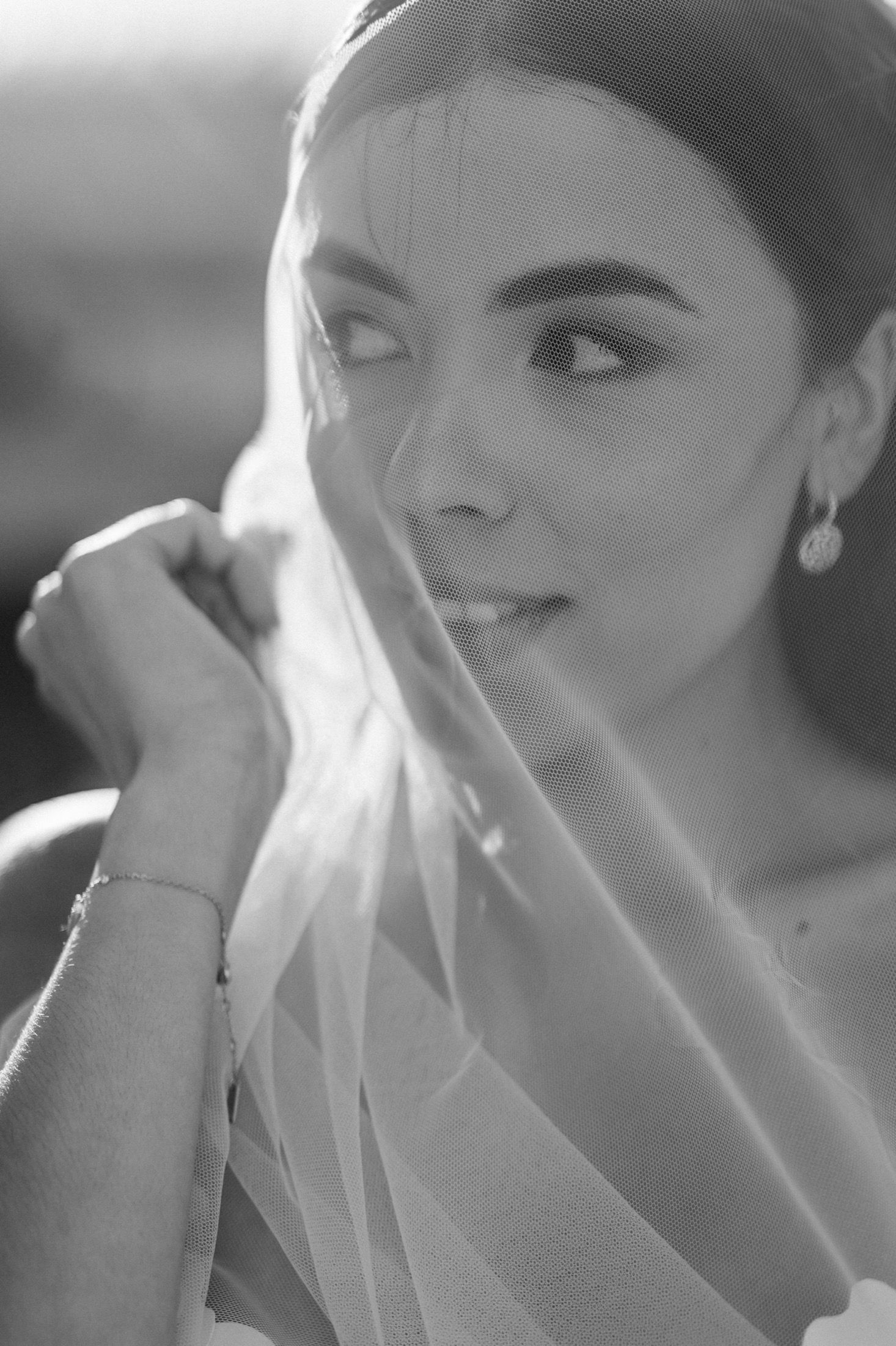 Black and white bridal portrait of a brunette bride holding her veil near her face, wearing pearl earrings and bracelet.
