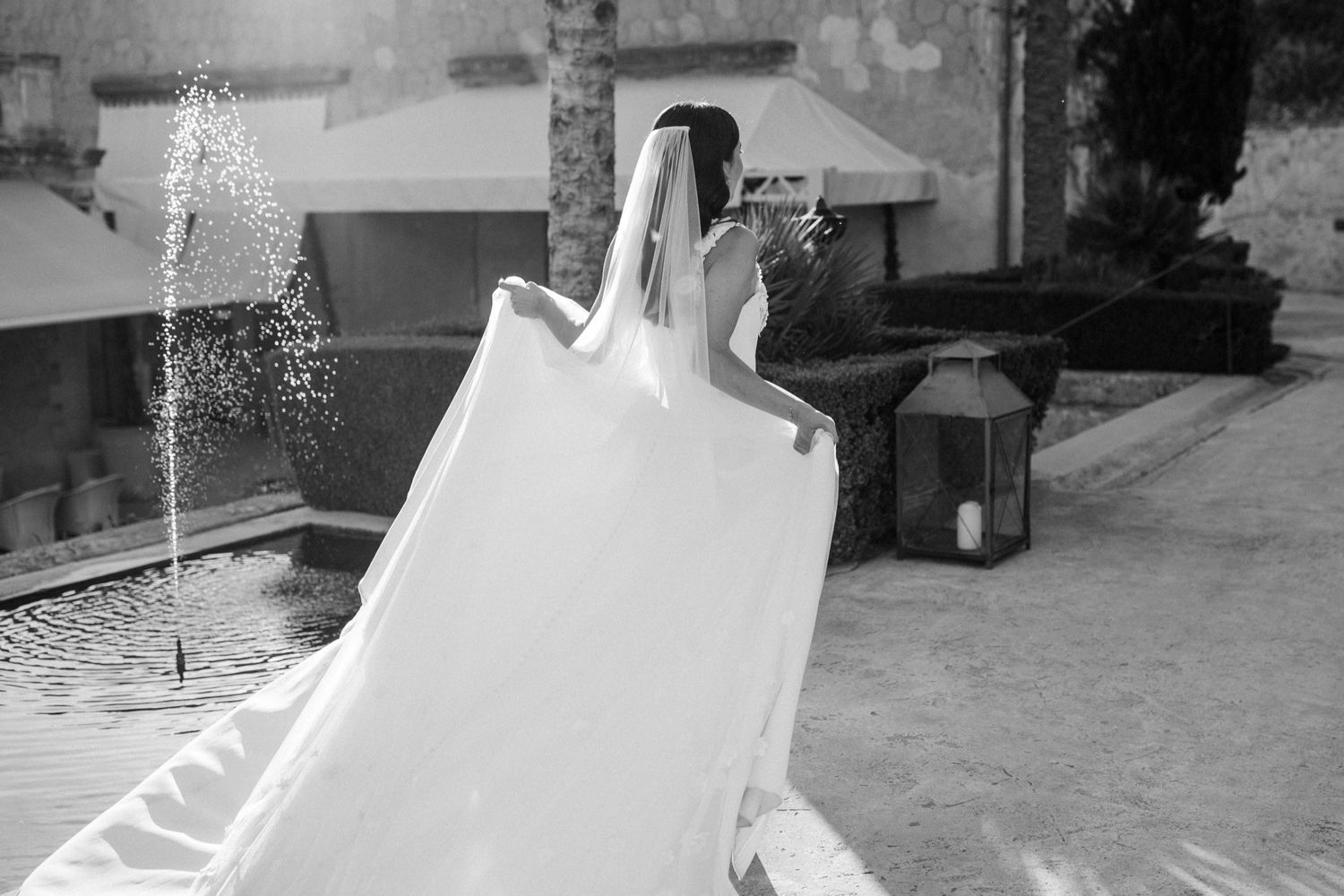 Black and white photo of a bride in a flowing wedding gown and veil near a fountain at an elegant outdoor venue.