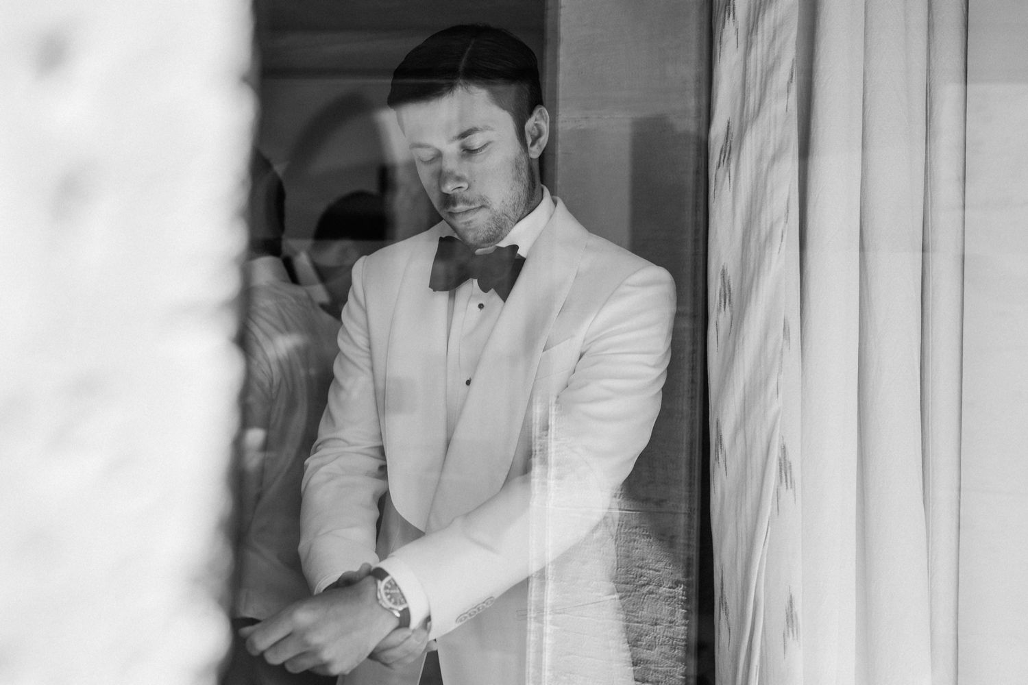 Black and white photo of a groom in white tuxedo and bow tie adjusting his cufflinks by a window.