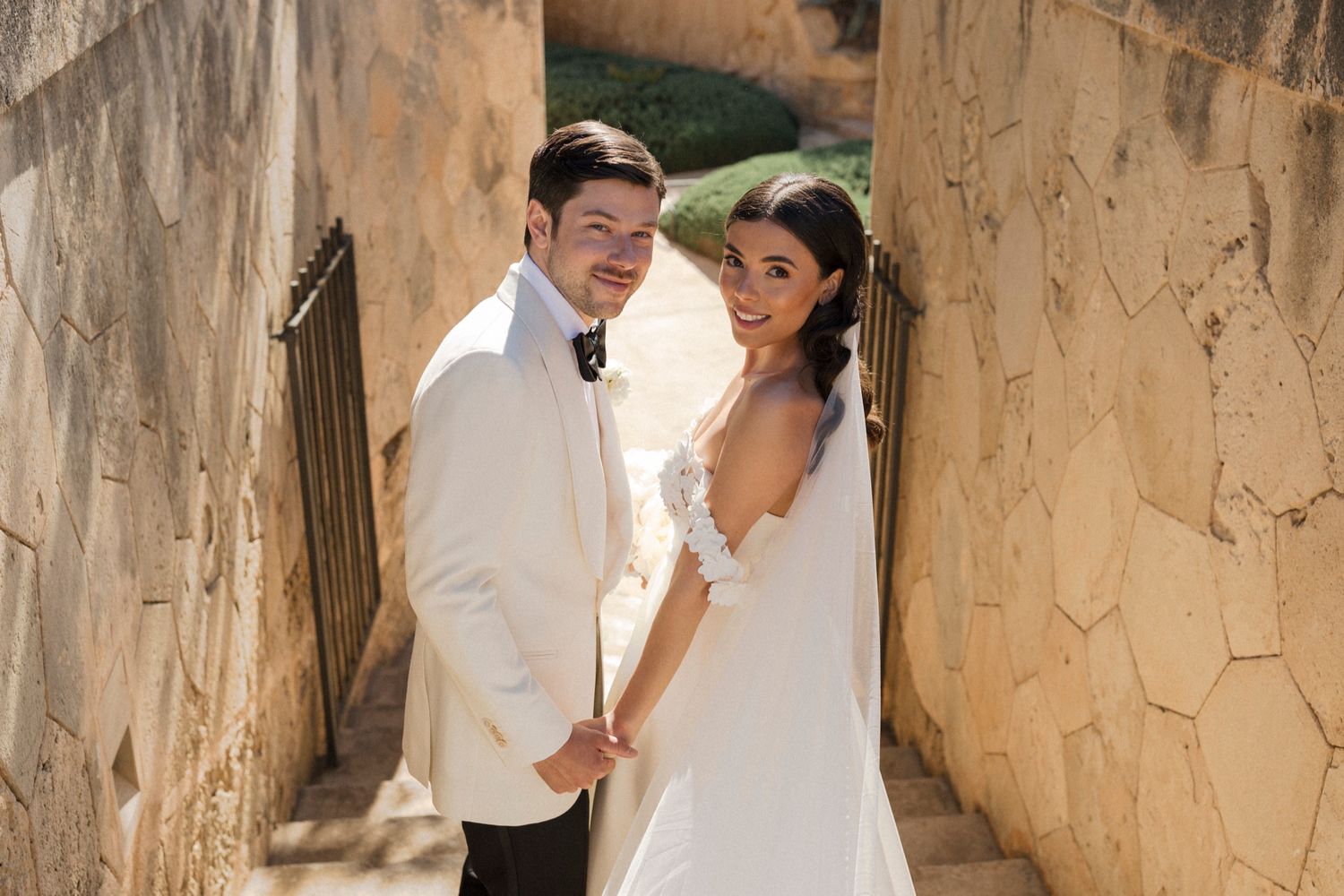Bride and groom holding hands in a sunlit stone alleyway, smiling at the camera on their wedding day.