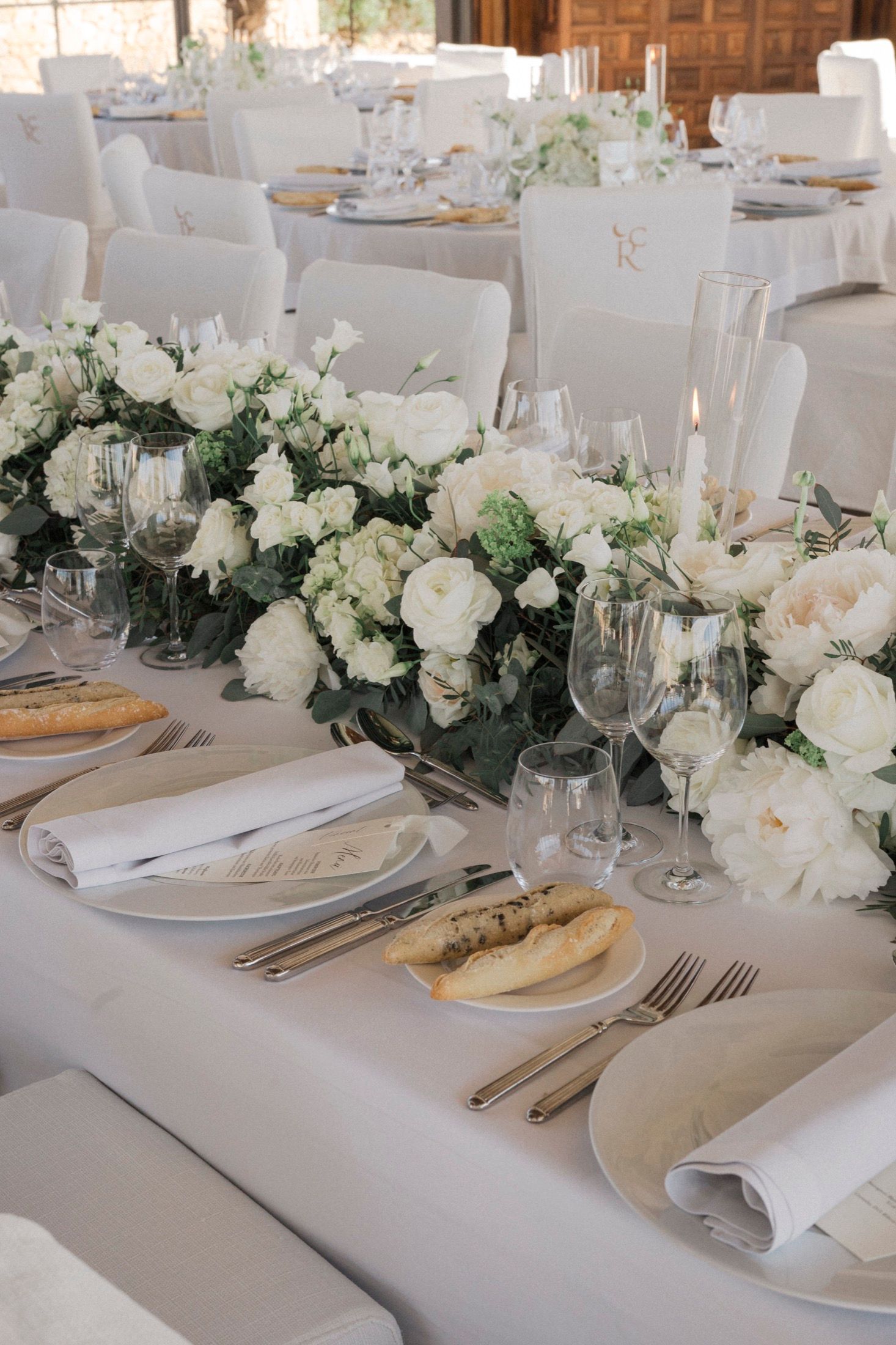 Elegant wedding reception table with white floral centerpieces, fine china, crystal glasses, and gold cutlery on white linen.