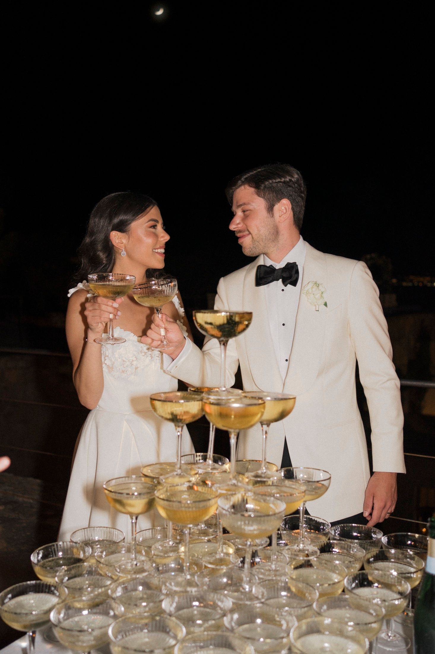 Bride and groom toasting with champagne coupe glasses beside a elegant champagne tower at their wedding reception.