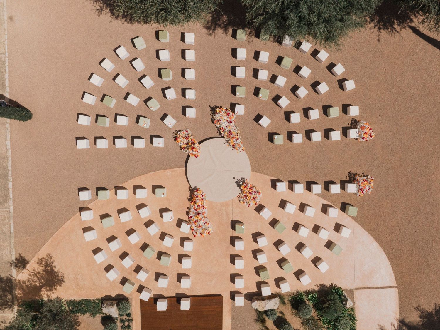 Aerial view of white beach chairs arranged in a circular pattern on sandy ground surrounded by green trees.