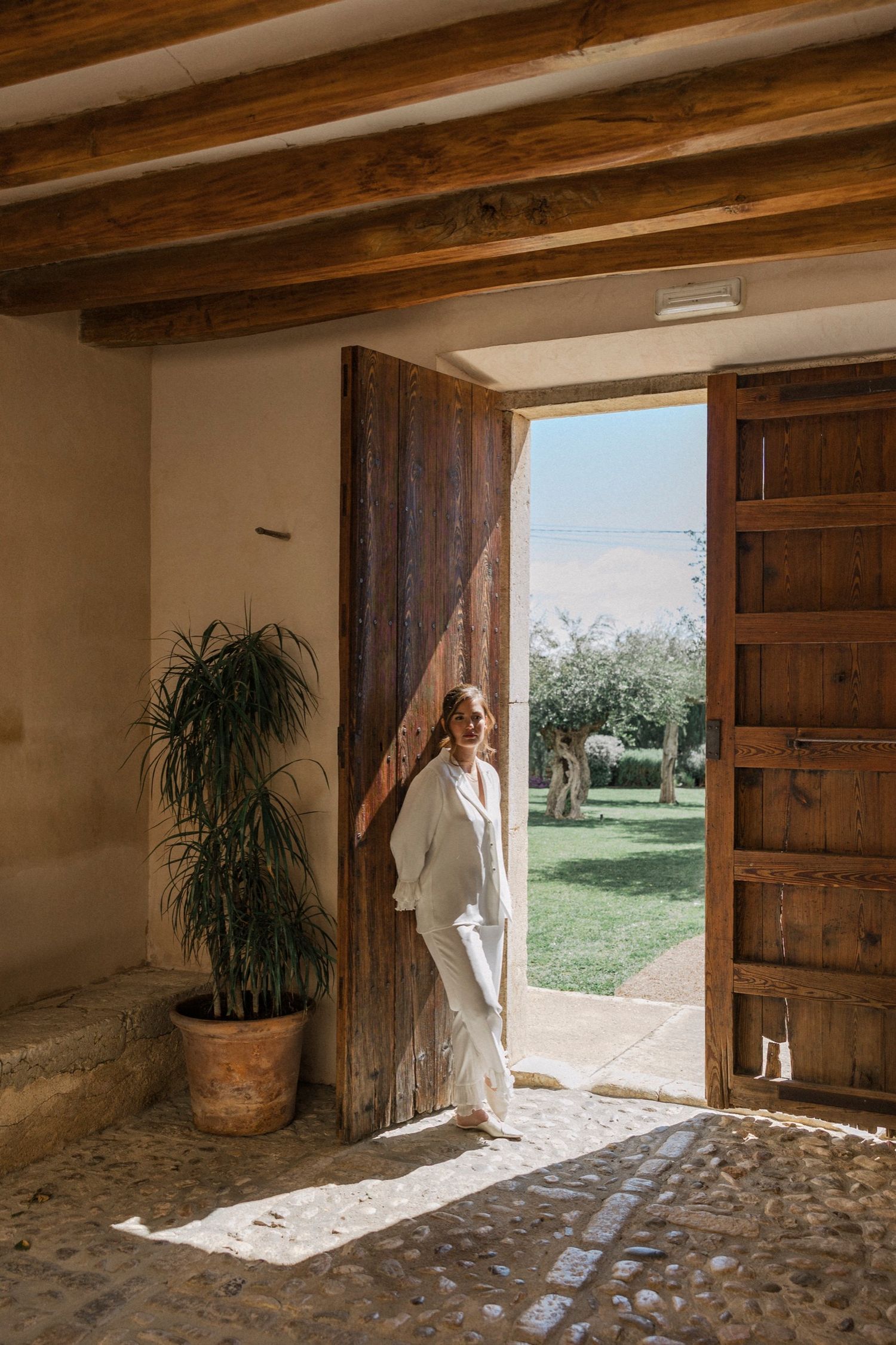 Sunlight streams through rustic wooden doorways into a Mediterranean style stone interior with potted plants.