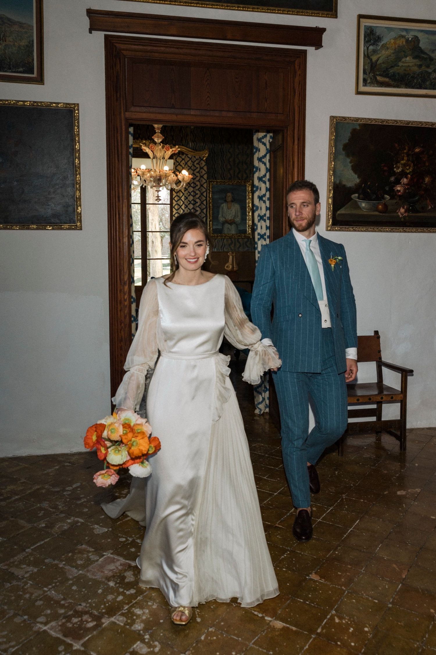 A couple walks through an ornate hallway with stone floors and artwork on walls during their wedding celebration.