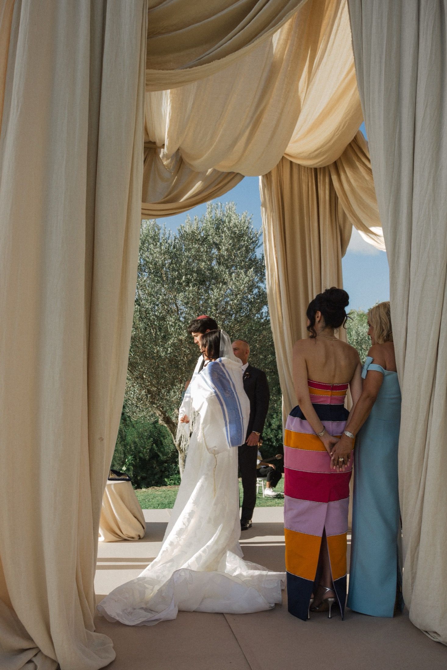 Wedding party gathers under elegant draped white curtains in an outdoor venue with olive trees in the background.