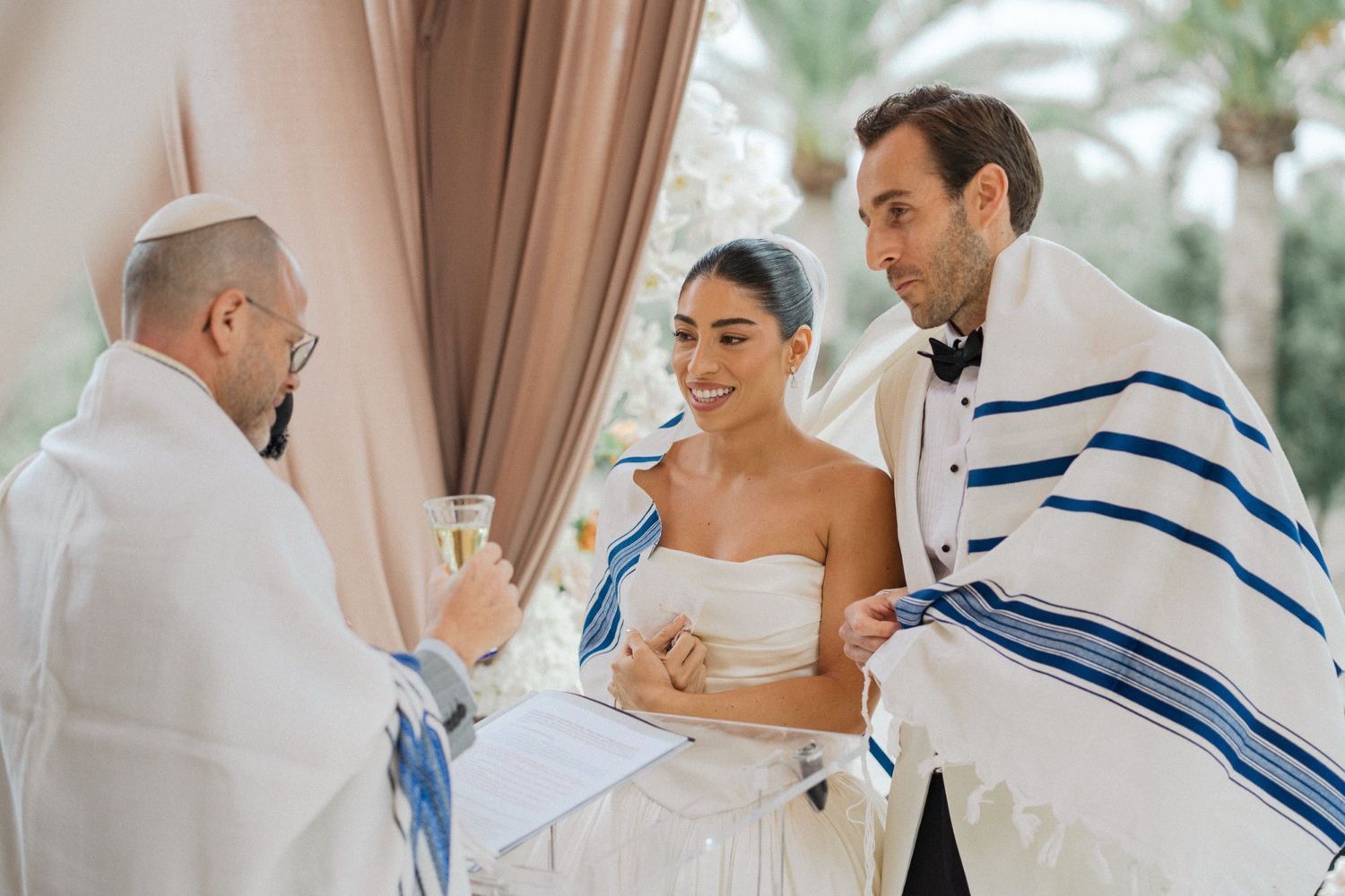 A bride and groom share a joyful moment during their Jewish wedding ceremony under a traditional prayer shawl.