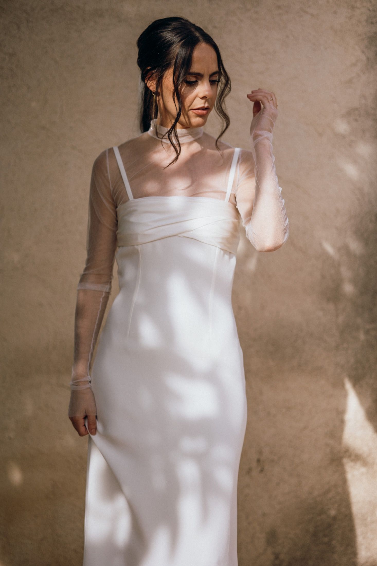 A person wearing a simple white slip dress poses in soft natural lighting against a textured beige wall.