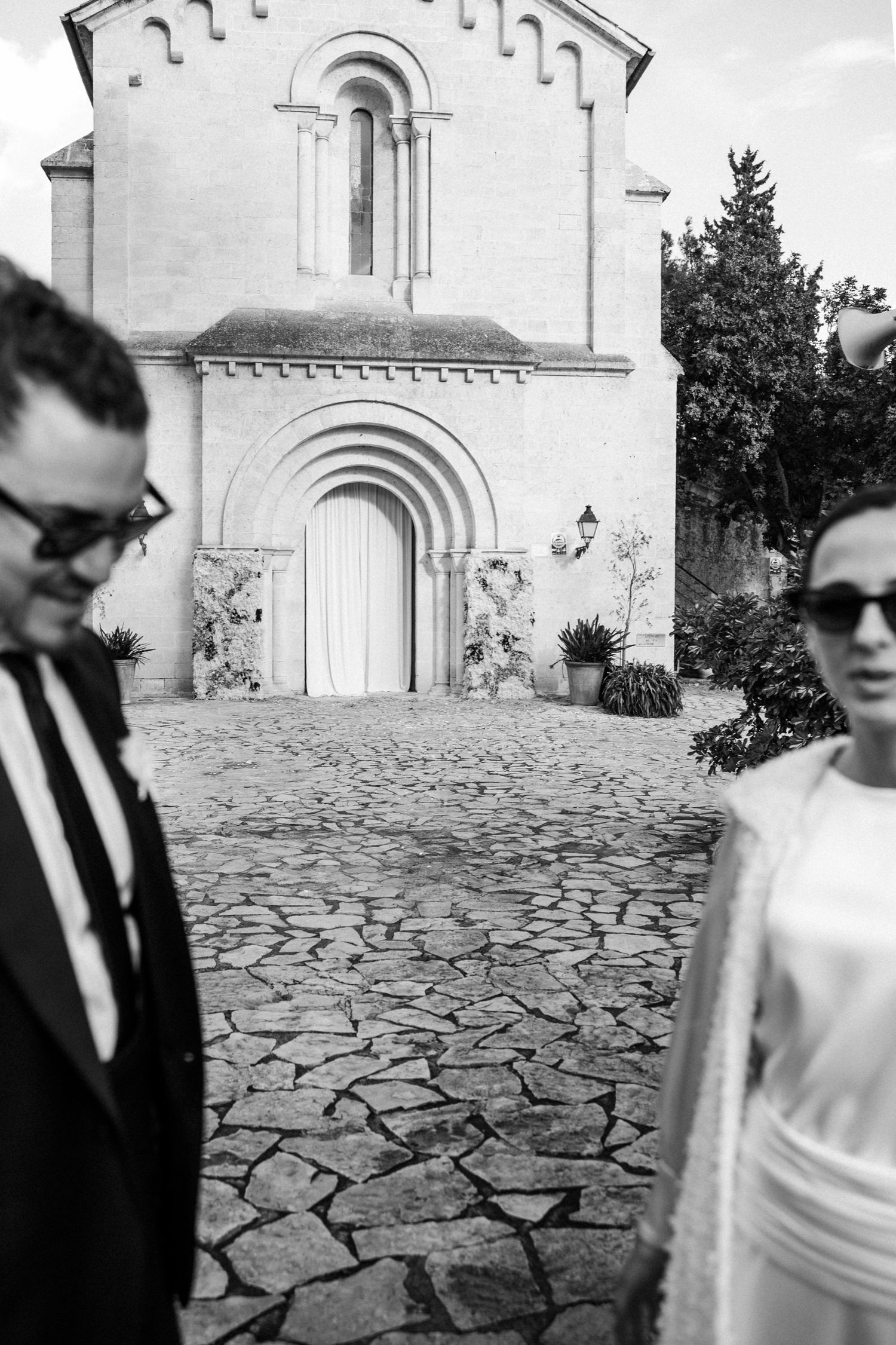 Elegant couple in formal wear standing on cobblestone path in front of a historic church.