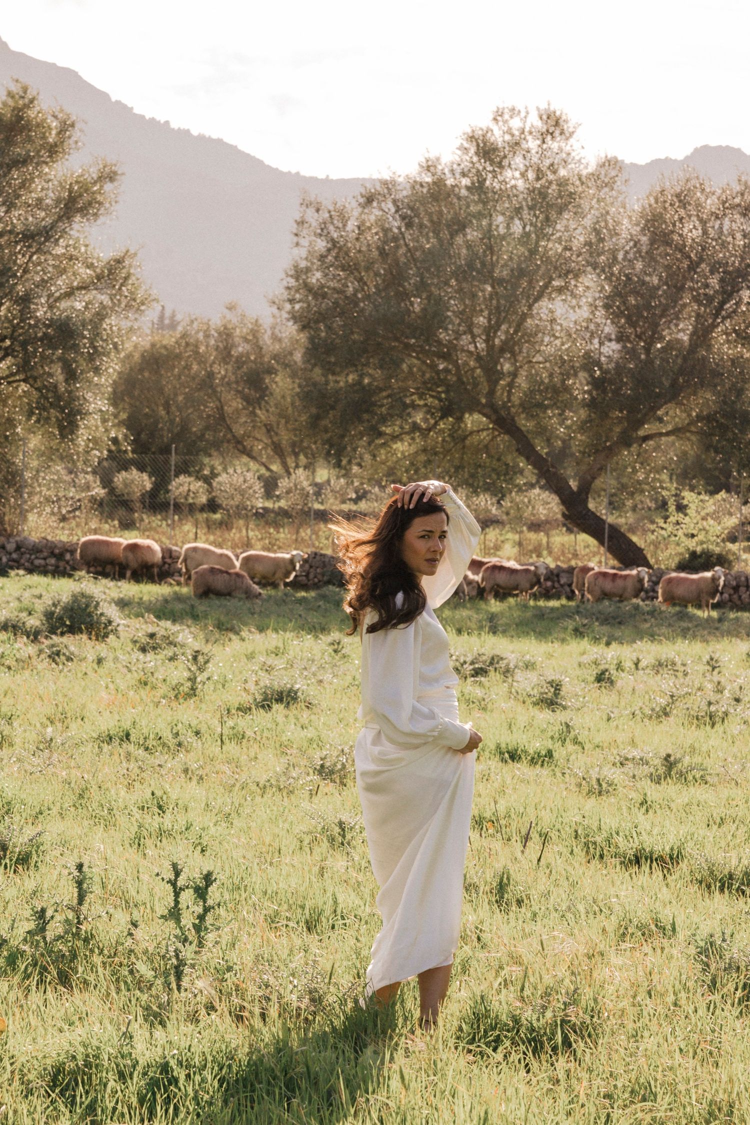 Person in white dress twirls in a meadow with sheep grazing nearby.