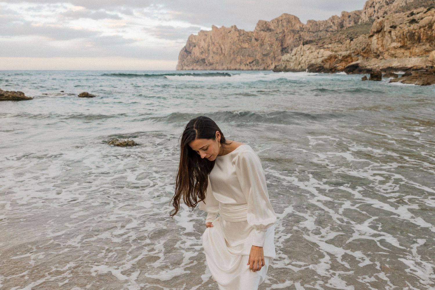 Woman in white outfit plays in ocean waves with dramatic coastal cliffs behind.