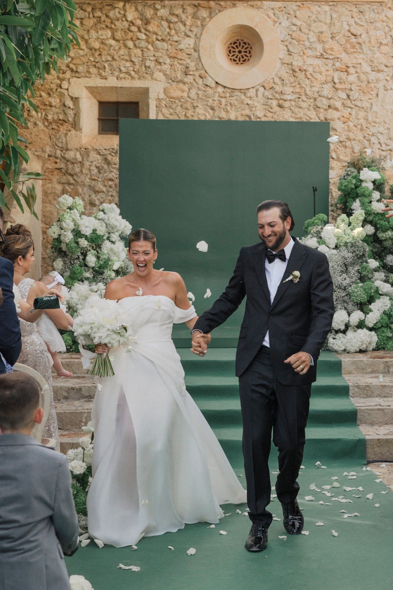 Wedding celebration moment on green floor with white floral decorations against stone wall.