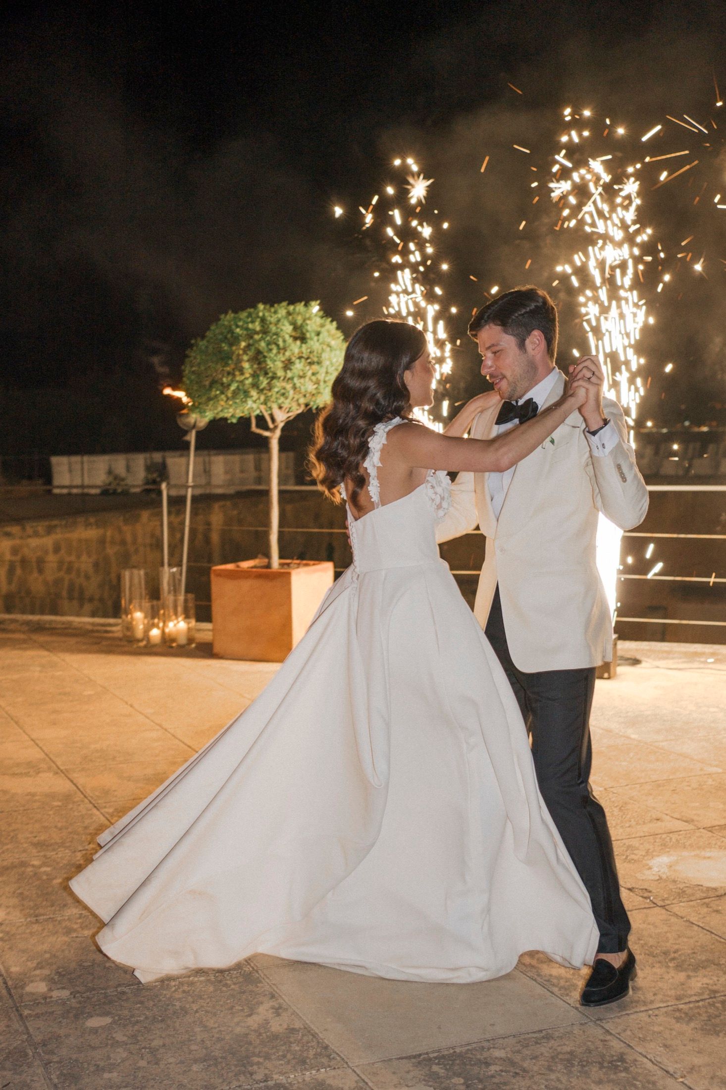 Bride and groom share first dance outdoors at night with sparkling fireworks fountain backdrop at elegant wedding reception.