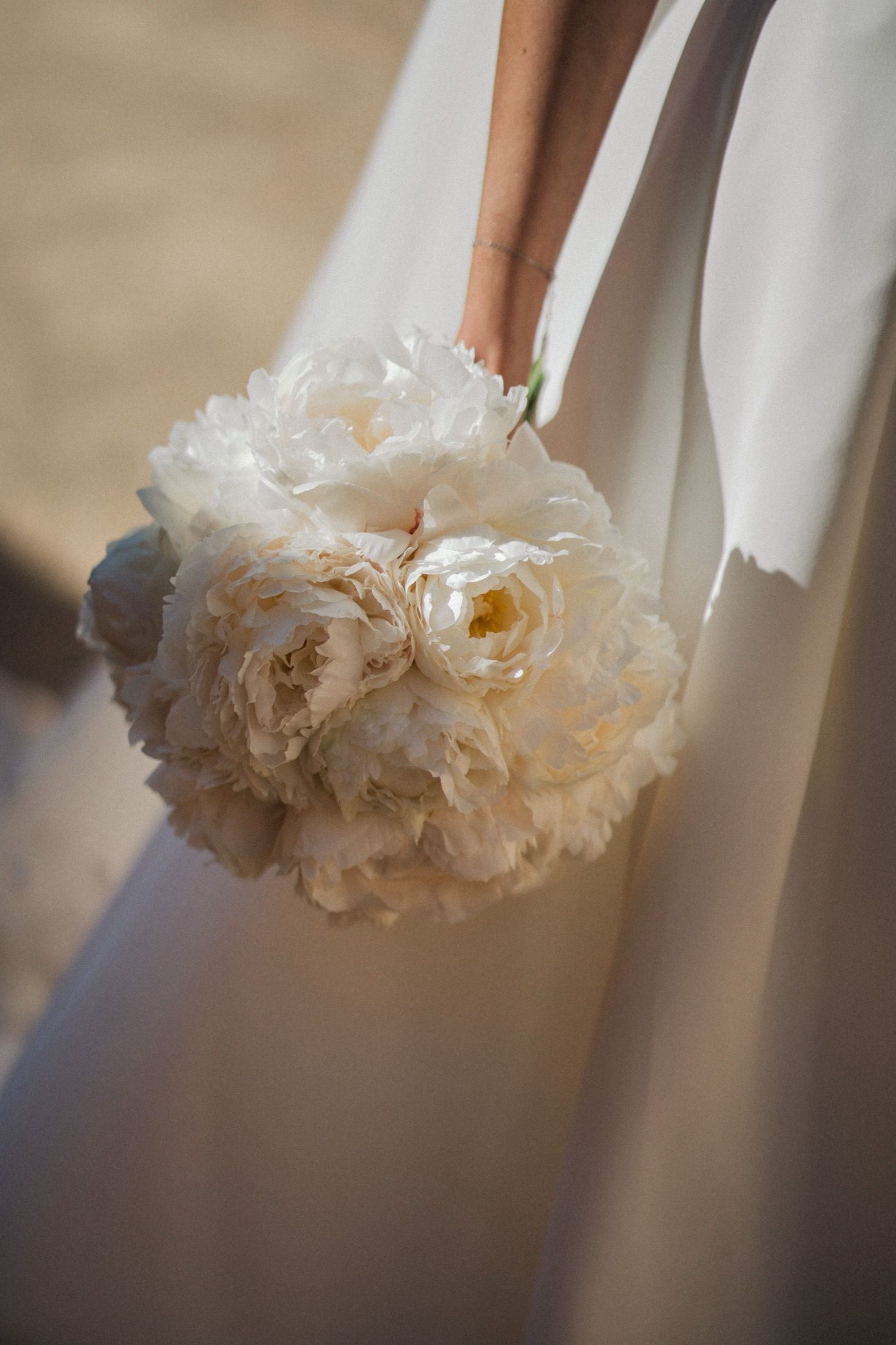 Bride holding a round white peony wedding bouquet against her flowing white gown.