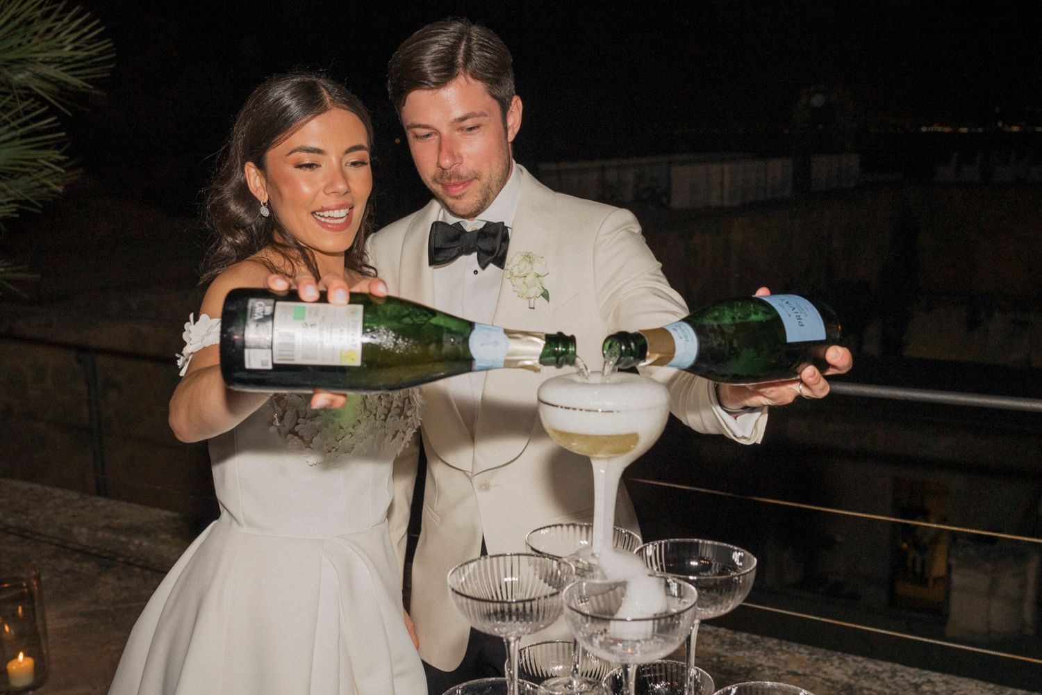 Newlyweds pouring champagne into a tower of glasses outdoors at night during their wedding celebration.