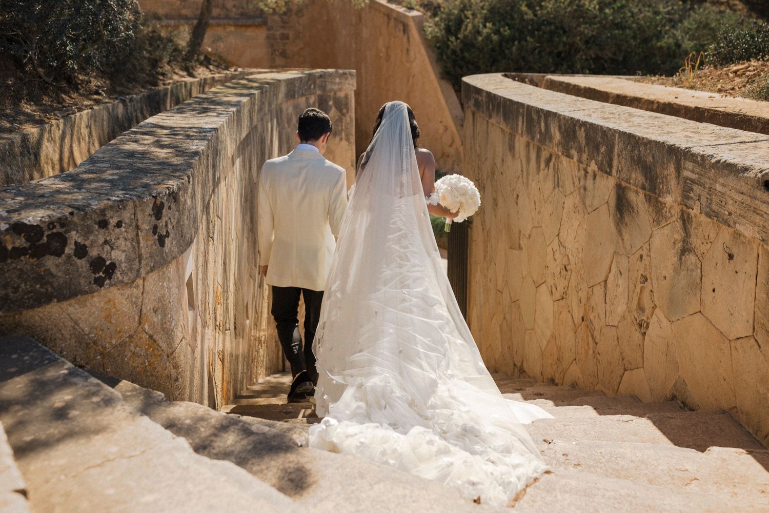 Bride and groom walking up stone steps, bride in white gown with long veil holding bouquet, groom in white suit.