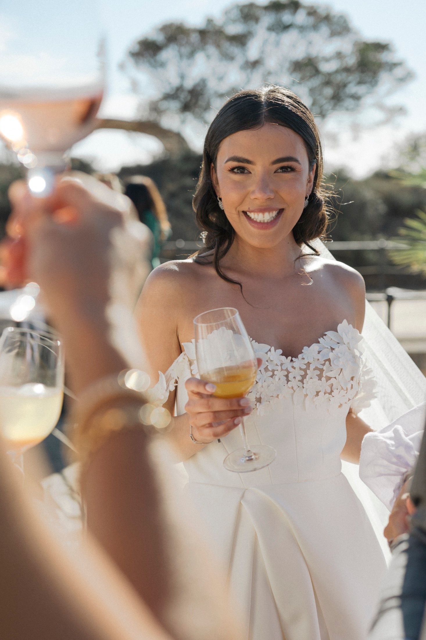 Smiling bride in off-shoulder lace wedding gown holding a champagne glass during an outdoor wedding reception.