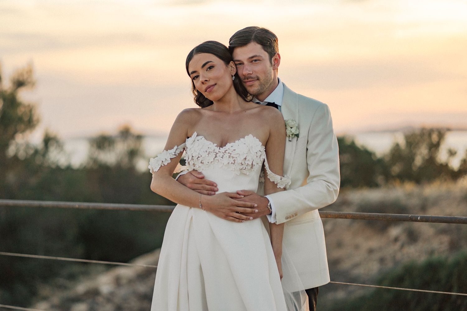 Bride and groom embrace at sunset, she in a lace wedding gown, he in a beige suit, with a scenic coastal backdrop.