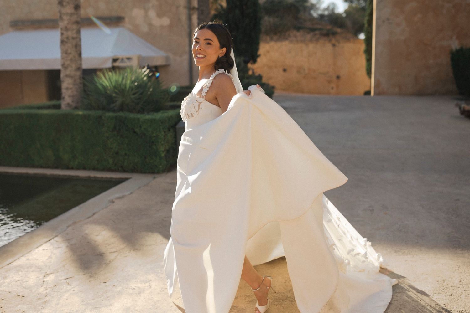 Smiling bride in elegant white gown lifting her dress outdoors near a garden.