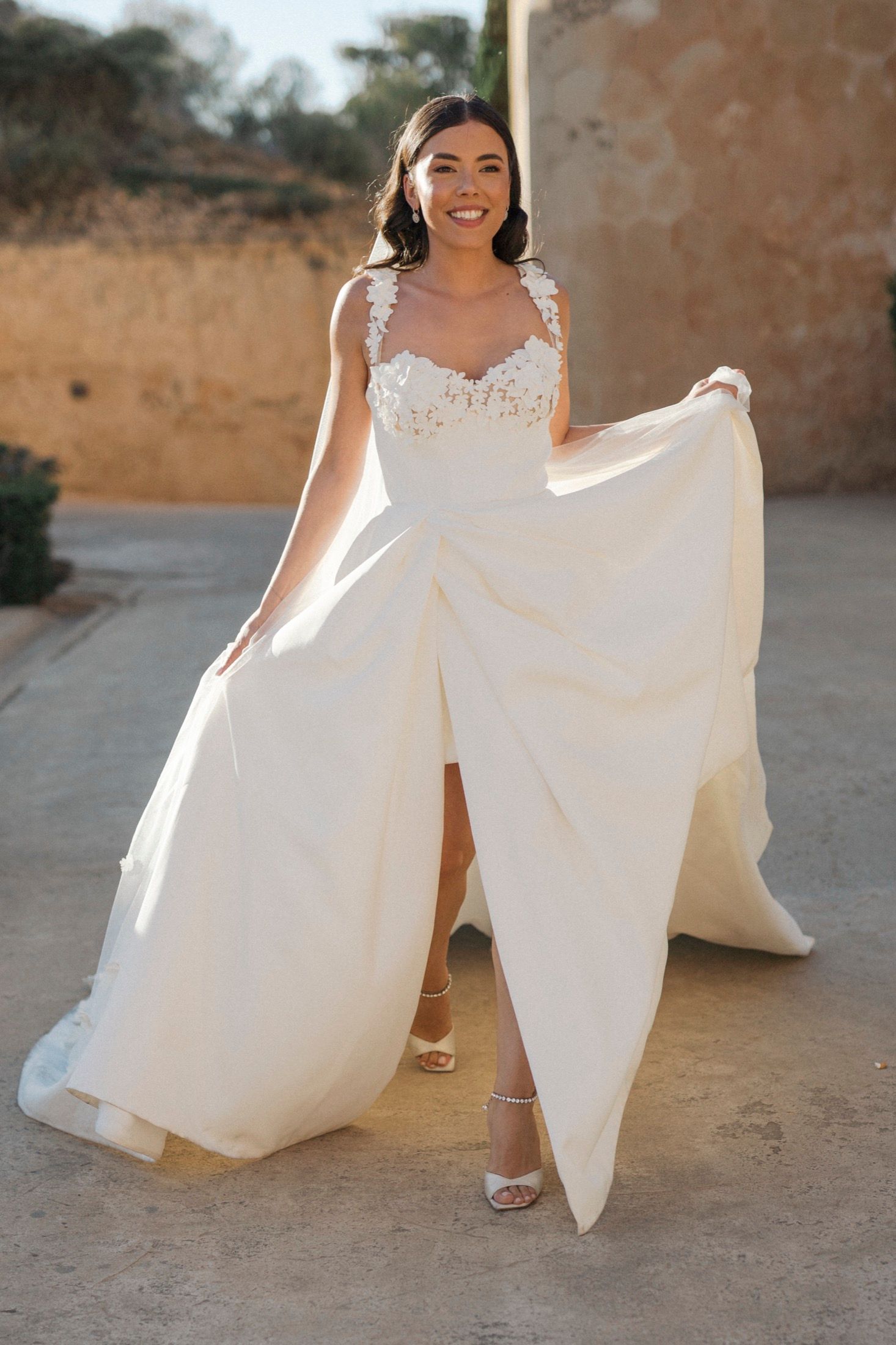Smiling bride in elegant ivory lace wedding gown with slit, holding skirt wide outdoors.
