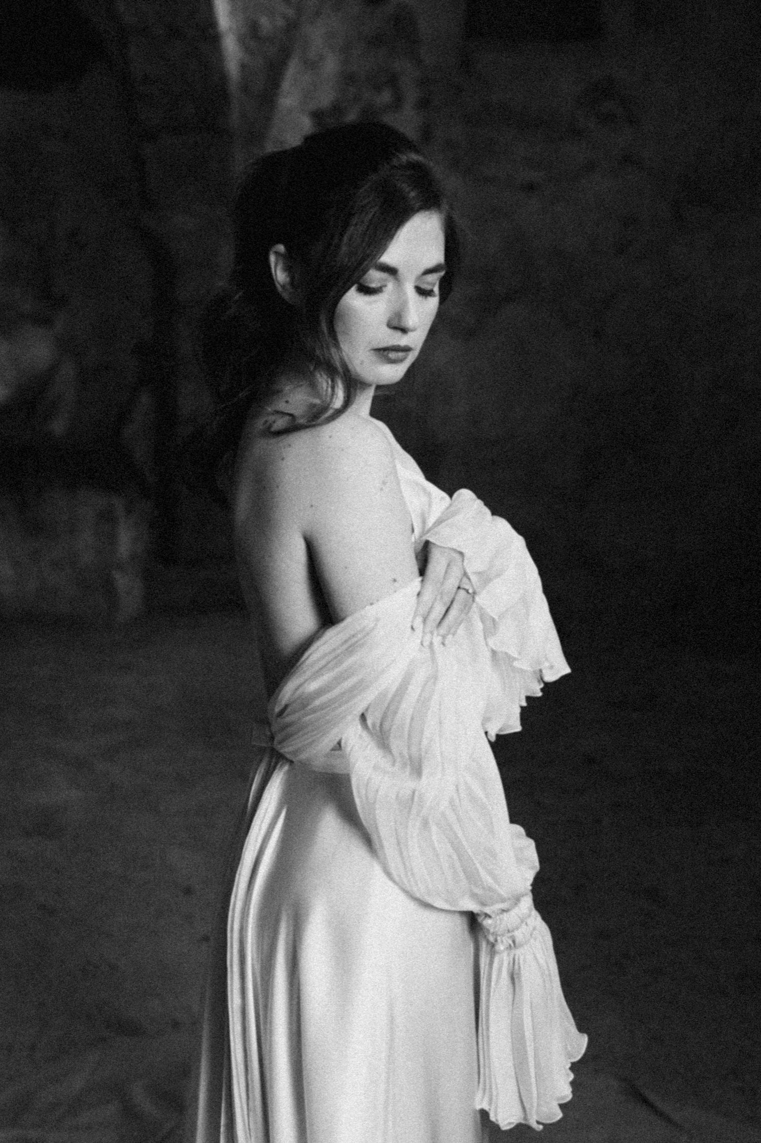 A dramatic black and white portrait of a figure in a flowing white dress with ruffled details against a dark background.