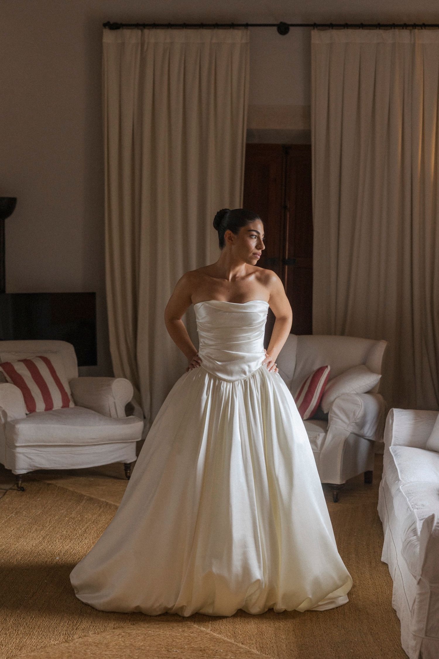 A bride in a white strapless wedding gown poses in a room with white slipcovered chairs and neutral decor.