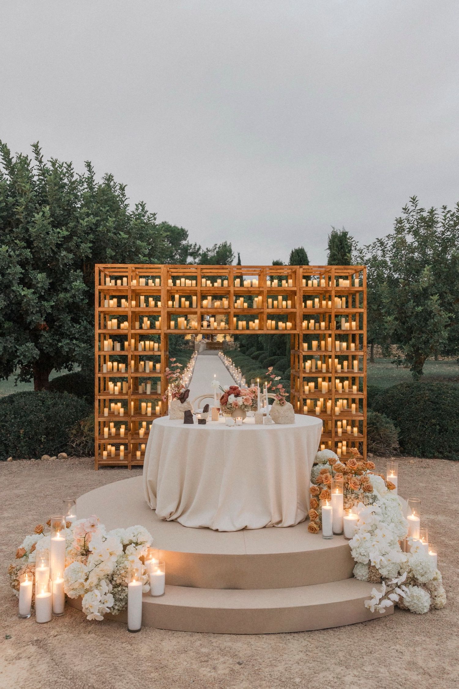 Elegant wedding reception setup with illuminated wooden crate backdrop, white floral arrangements and candles on curved platform.