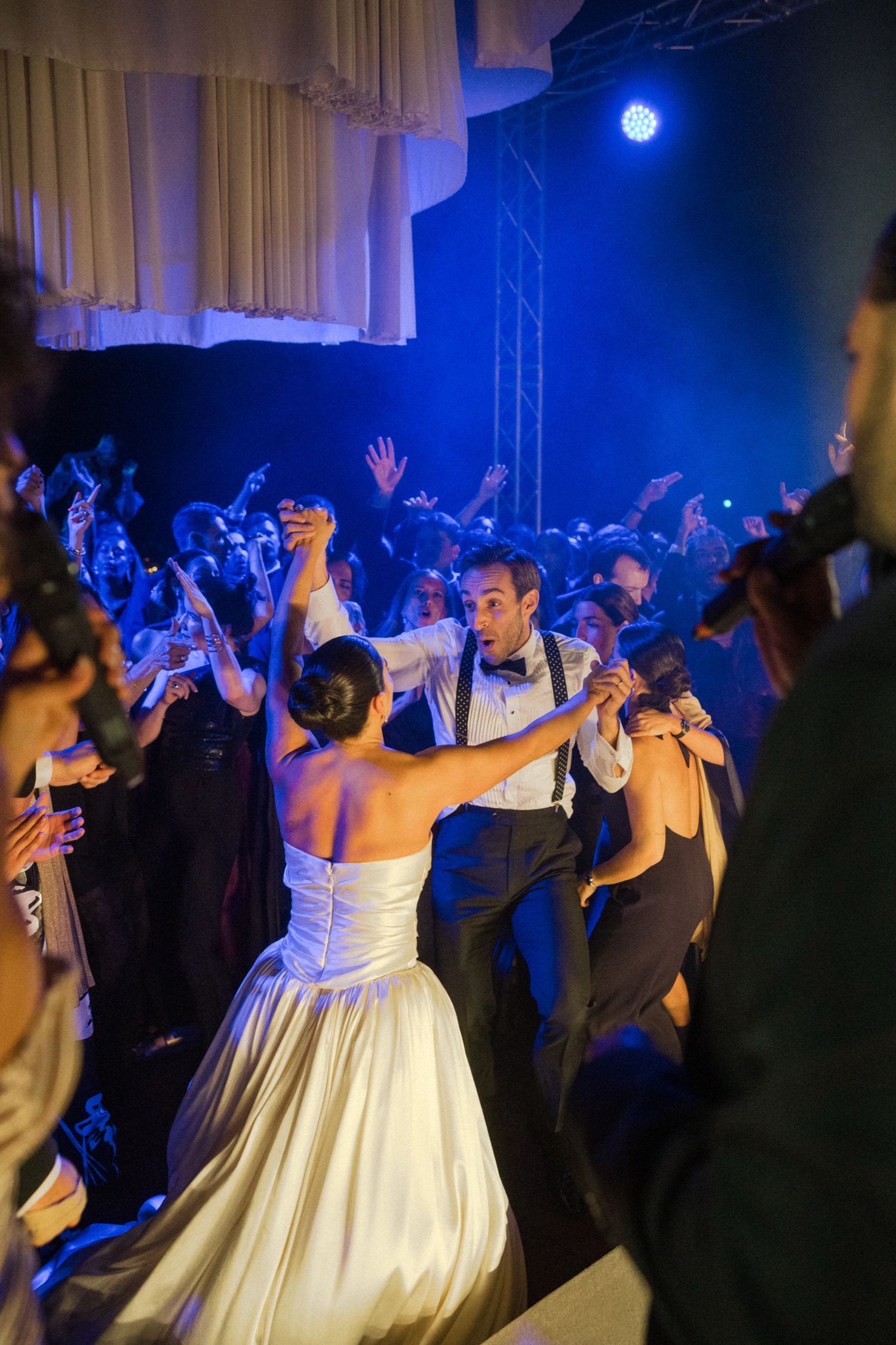Wedding guests dance and celebrate on a dimly lit dance floor illuminated by blue stage lighting at a reception party.