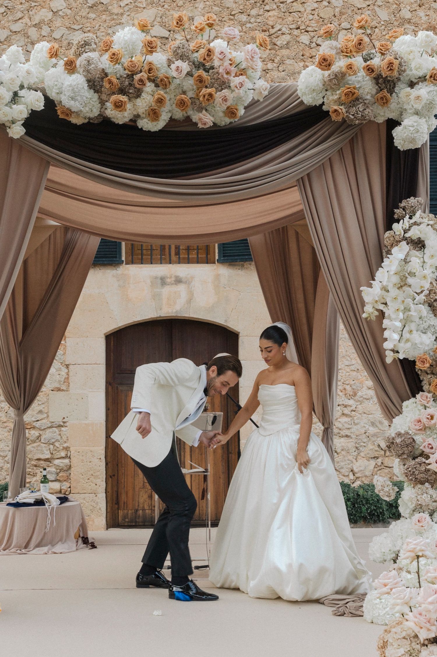 Wedding couple dancing under draped fabric and floral arch with blush and white flowers in stone courtyard.